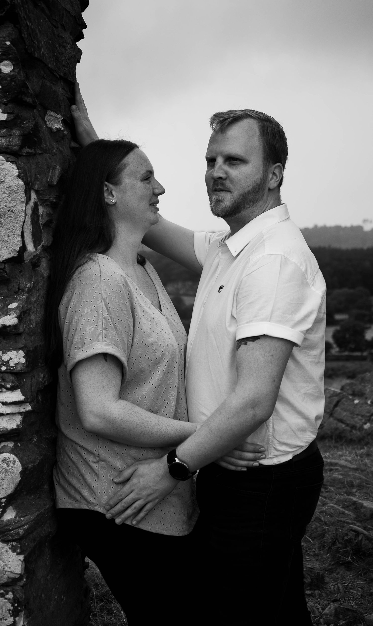 Black and white photo of a couple embracing outdoors near a stone wall, man tenderly holding womans face.