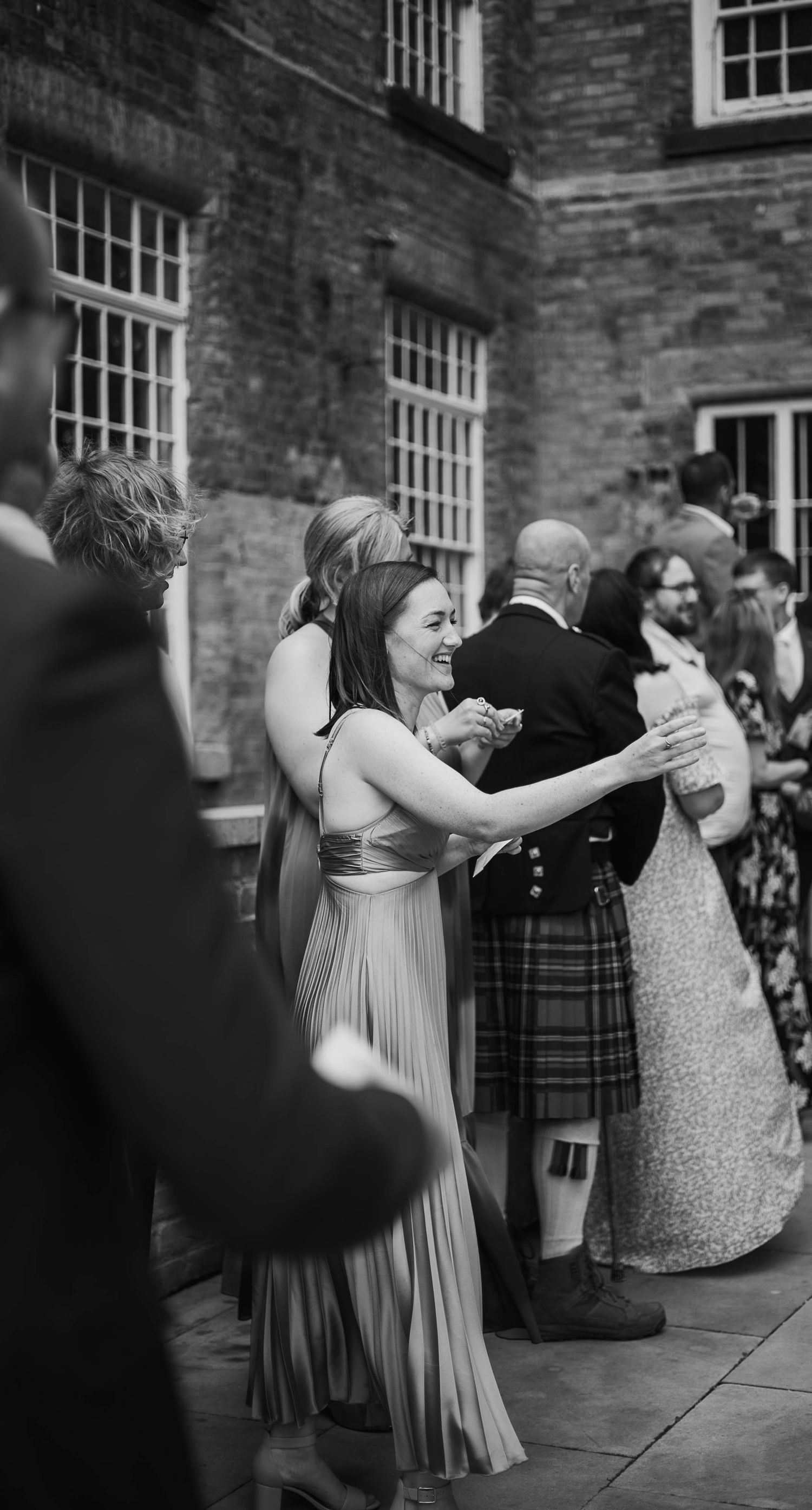 Guests dancing outdoors at a wedding reception, woman in elegant gown with man in traditional Scottish kilt, brick building backdrop.