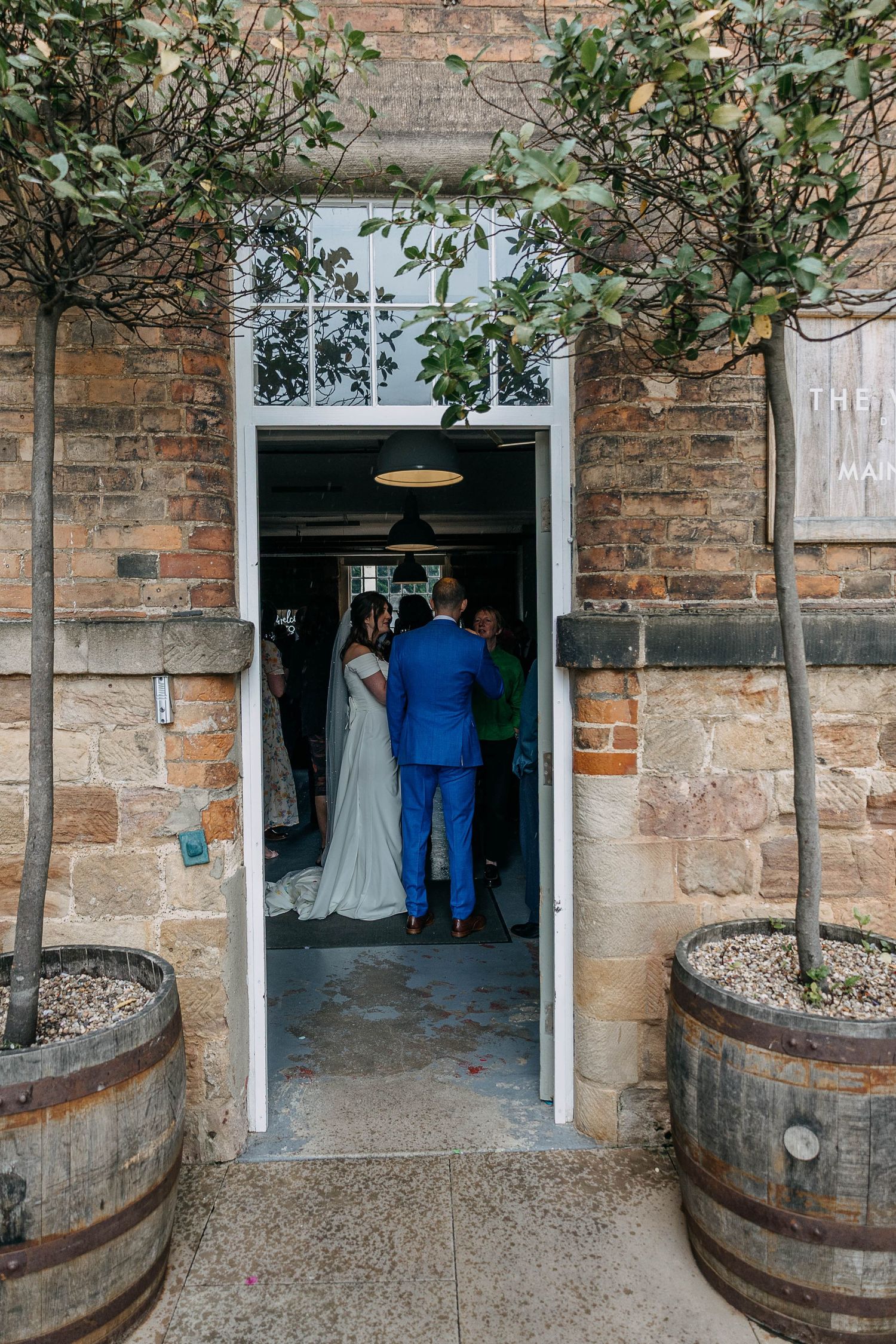 Bride and groom entering rustic stone wedding venue flanked by potted topiary trees and wooden barrels.
