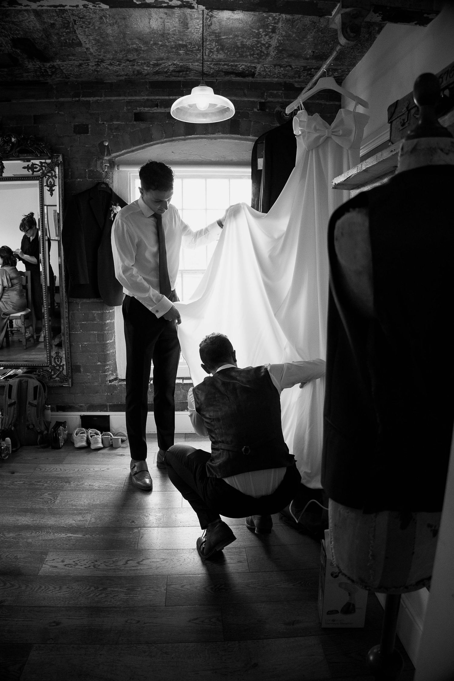 Black and white photo of a groom adjusting his tie while a tailor kneels to hem a wedding dress in a rustic room.