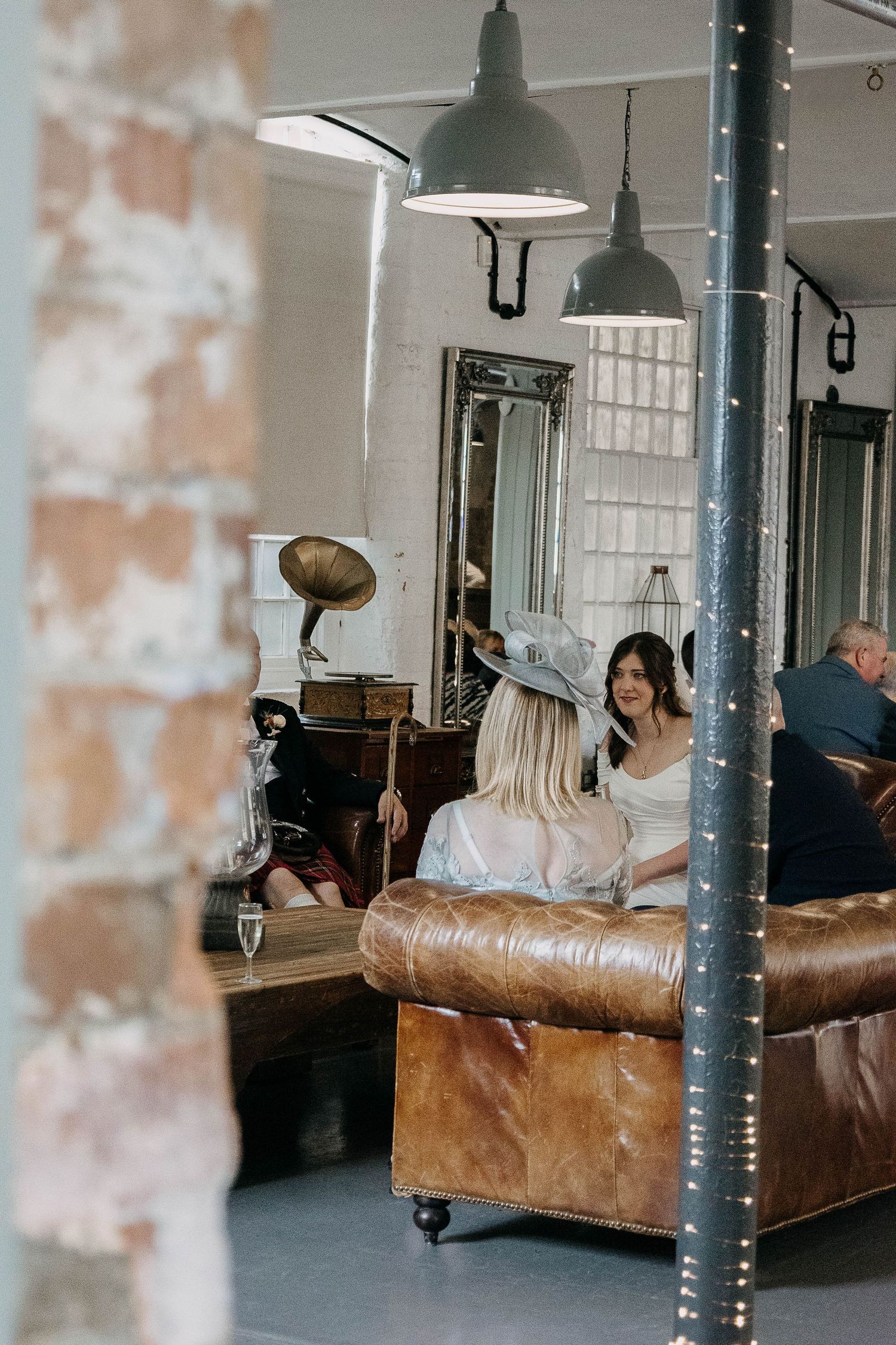 Two women chat on a leather sofa in a vintage industrial venue with pendant lights and a gramophone.