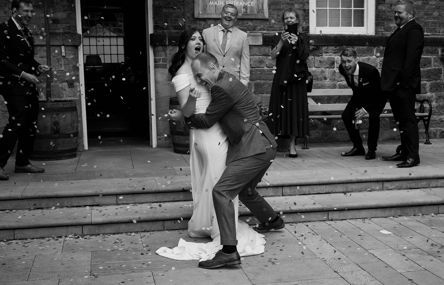Black and white wedding photo of groom lifting bride outdoors, guests celebrating with confetti at a brick building.