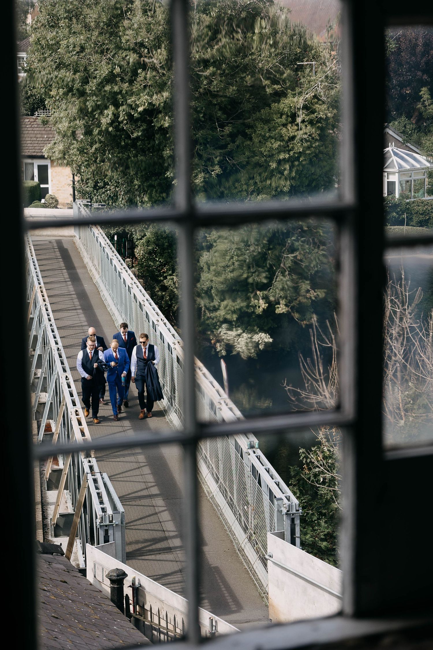 Wedding party in formal attire walking across a bridge, viewed through a leaded glass window.