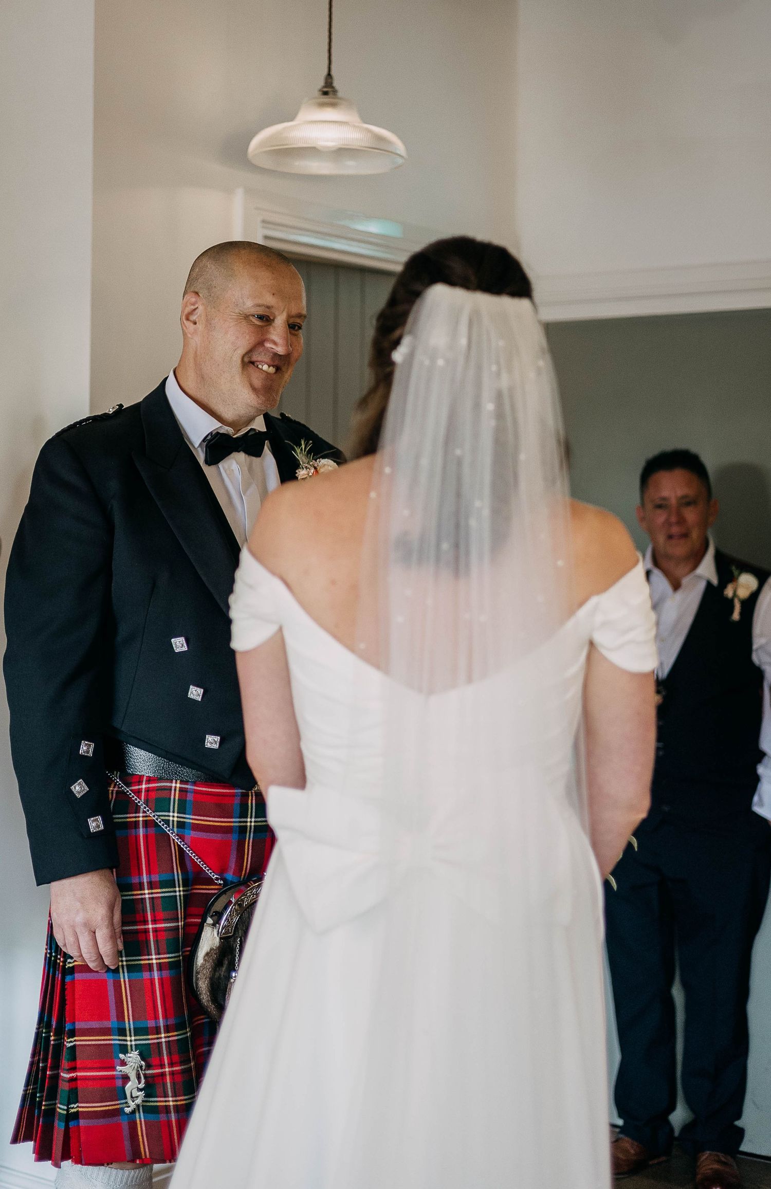 Groom in Scottish kilt smiling at bride in white wedding gown and veil during wedding ceremony.