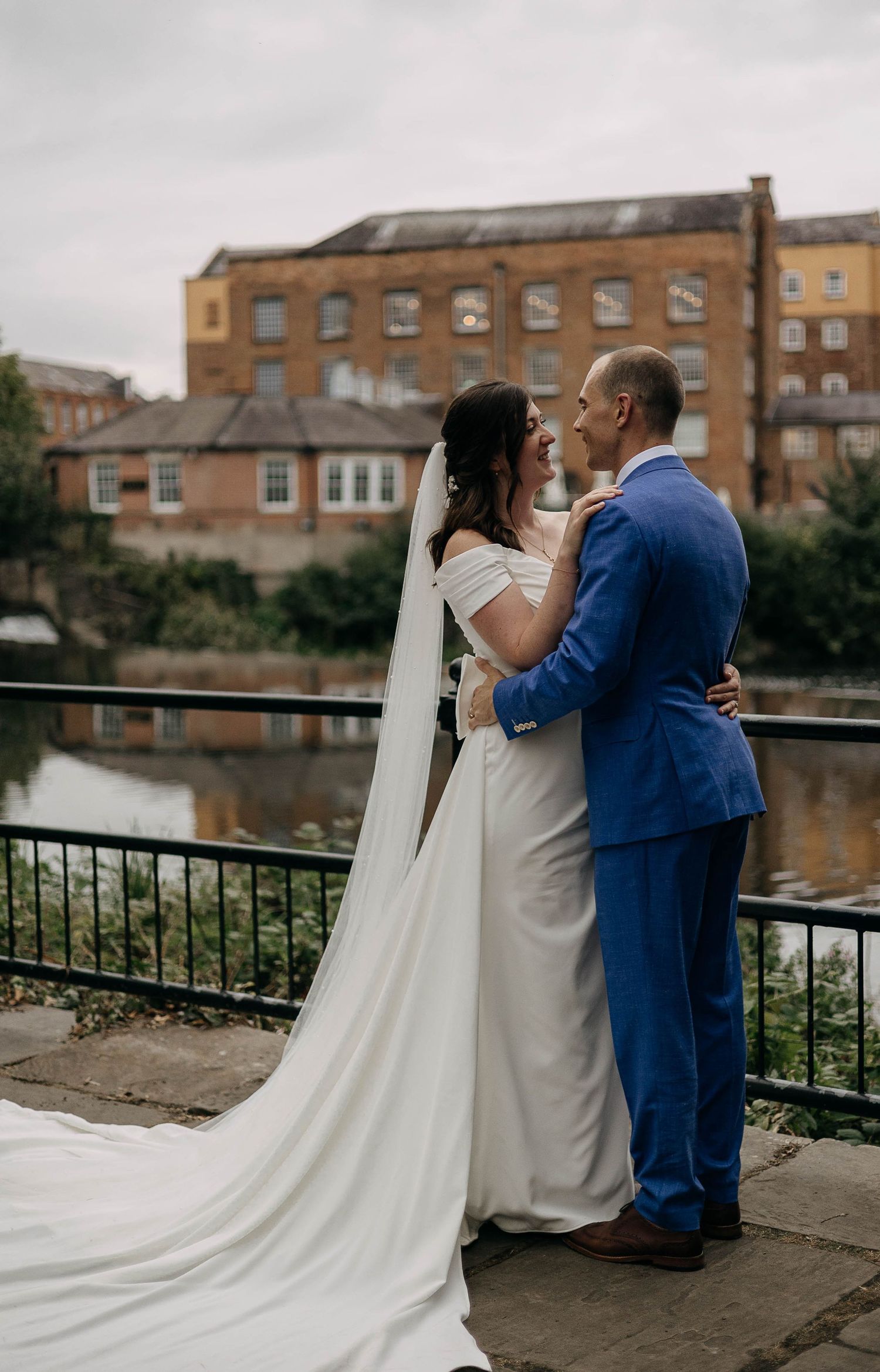 Bride and groom share a romantic moment by a riverside, wearing a white gown with veil and blue suit.