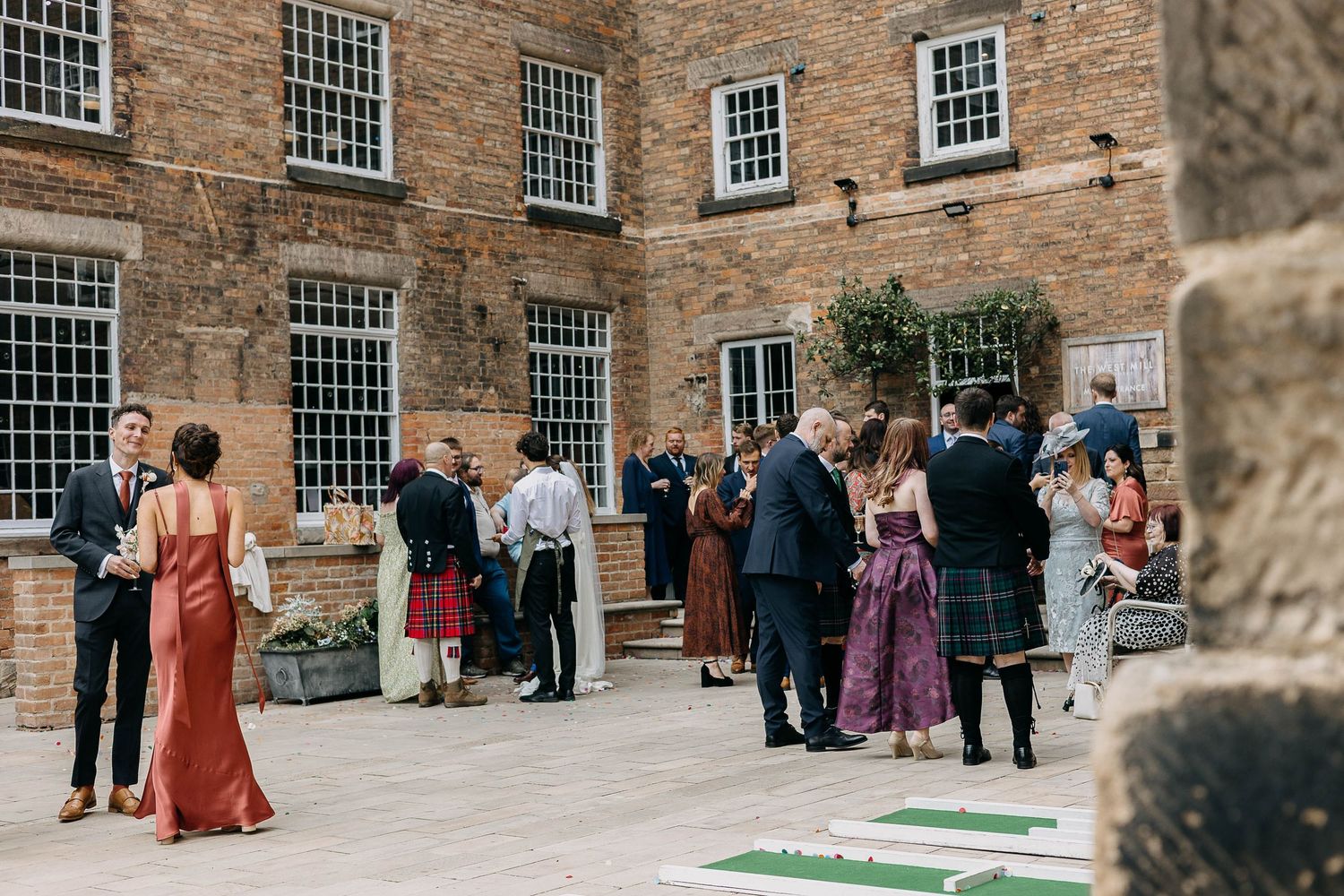 Guests mingle outdoors at a brick building wedding venue, some wearing kilts, enjoying a sunny garden reception.