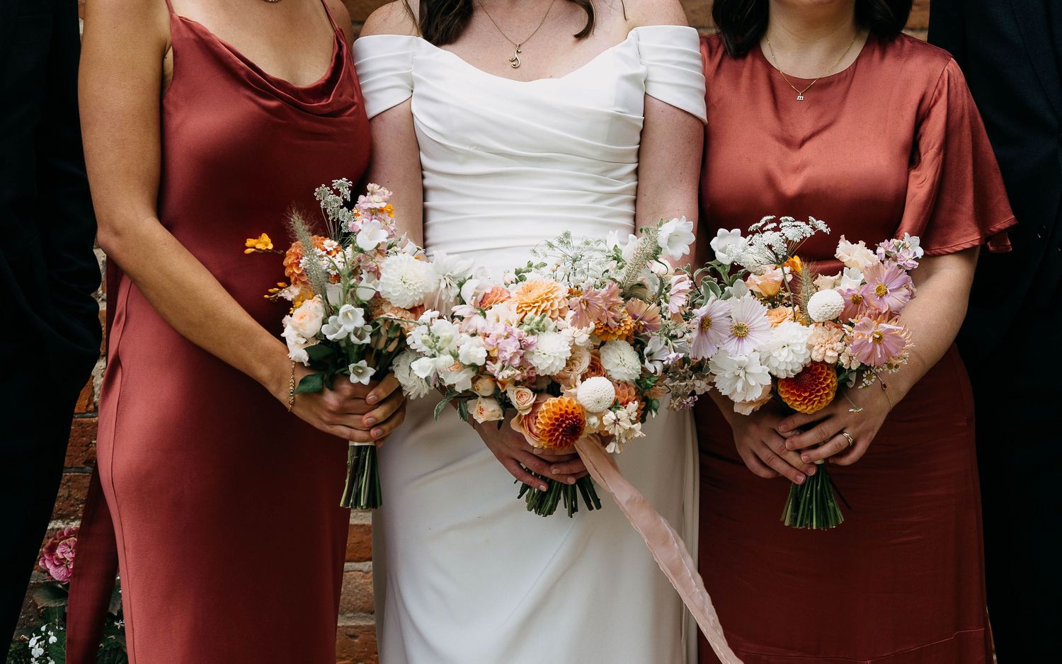 Bride in white off-shoulder gown flanked by bridesmaids in rust satin dresses, all holding lush floral bouquets.