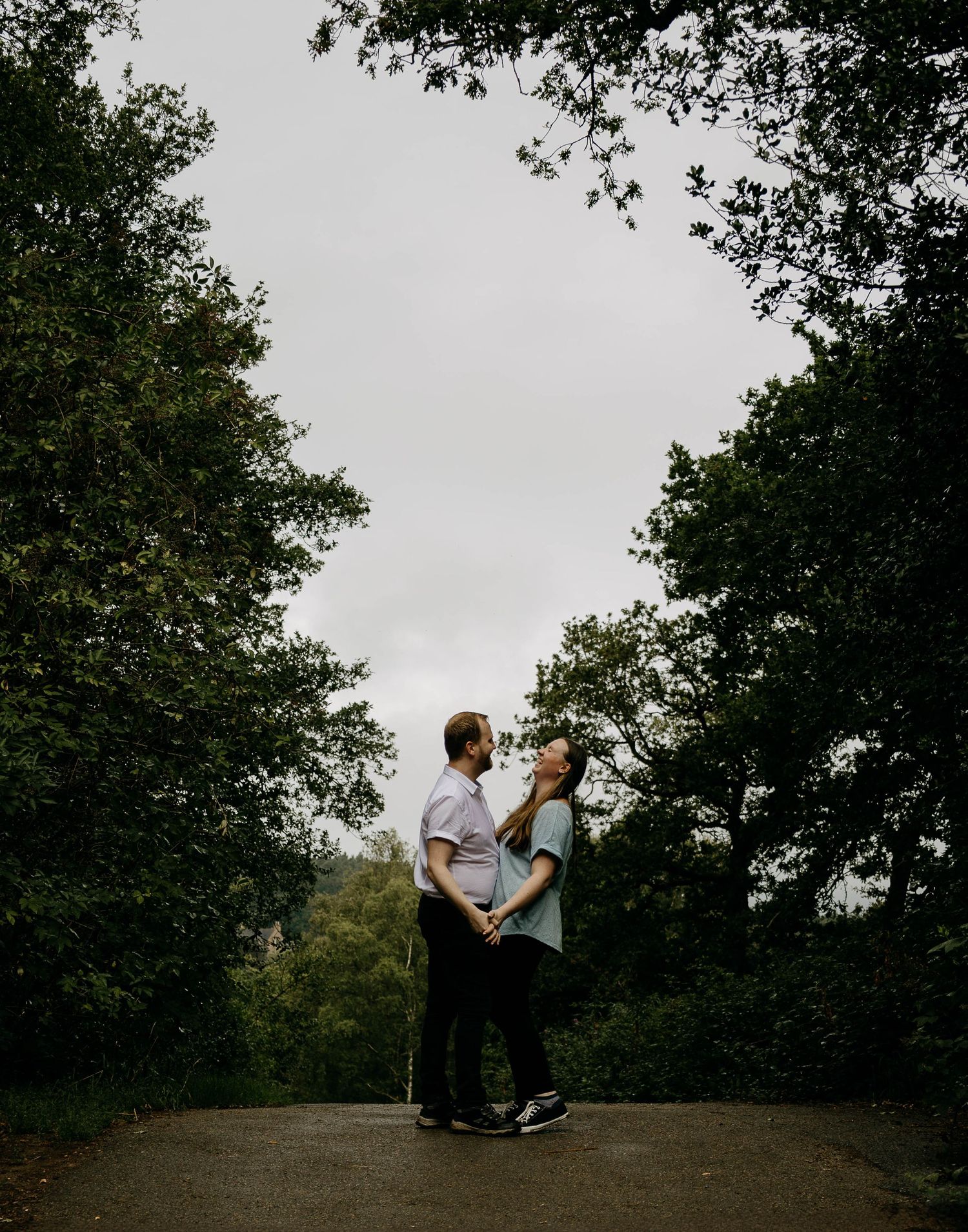 Couple standing together on a tree-lined path, sharing a tender moment under an overcast sky.
