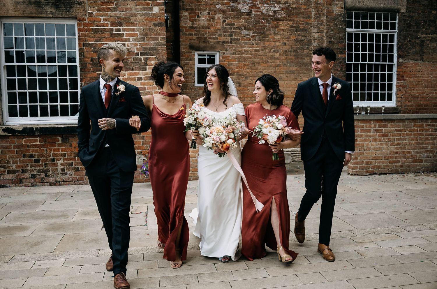 Bride in white gown walks with bridesmaids in rust dresses and groomsmen in navy suits at brick venue.