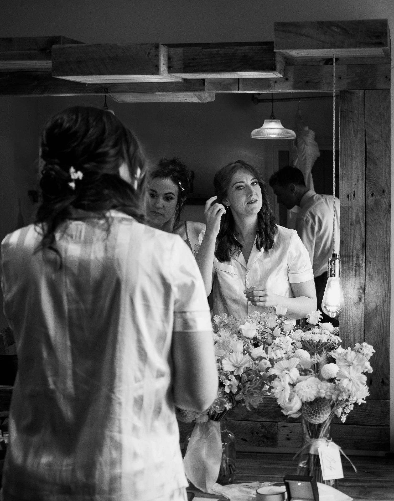Black and white photo of women getting ready, applying makeup near a mirror with flowers and rustic wooden decor.
