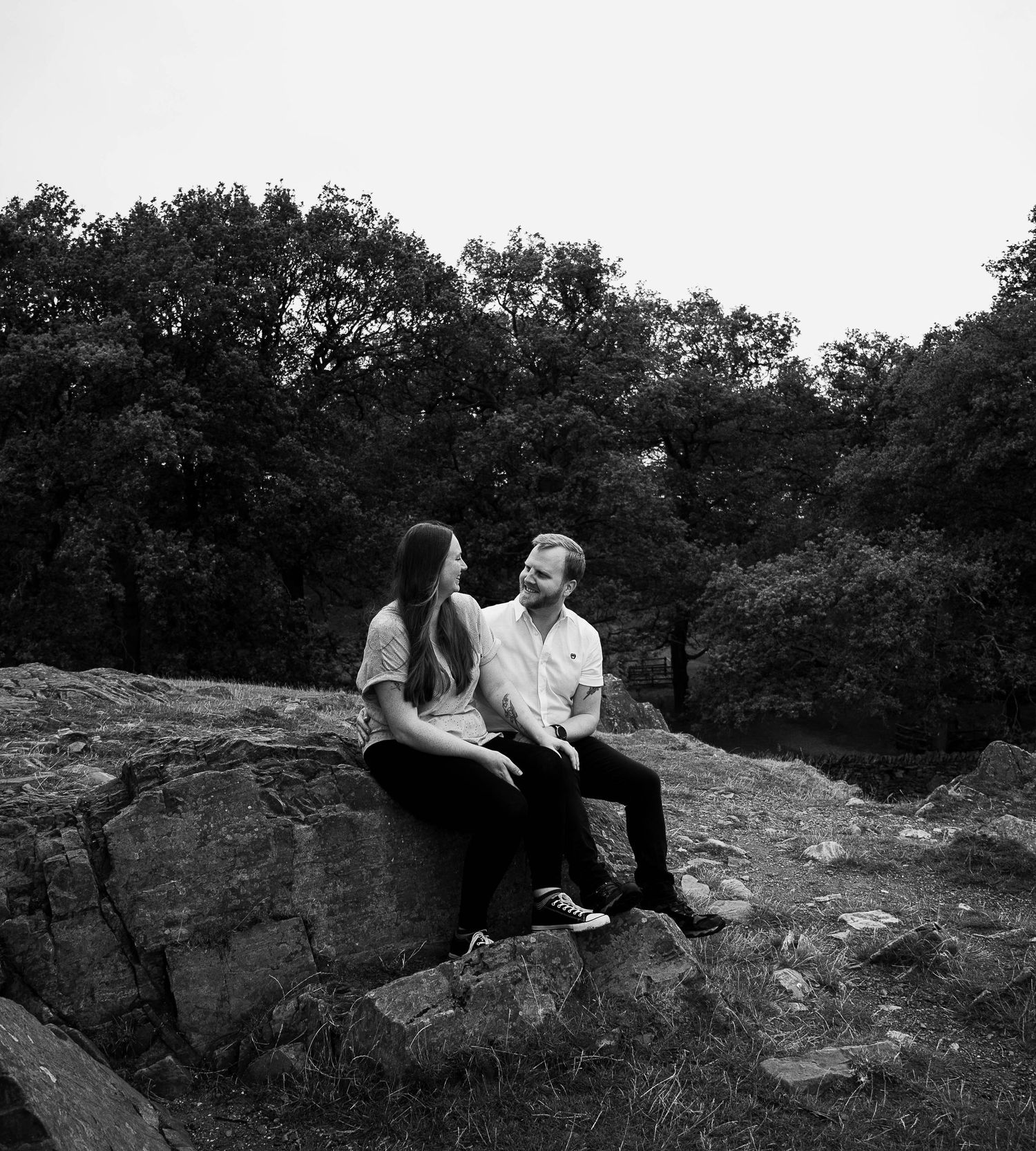 Black and white photo of a couple sitting on rocks surrounded by trees, sharing a quiet moment outdoors.