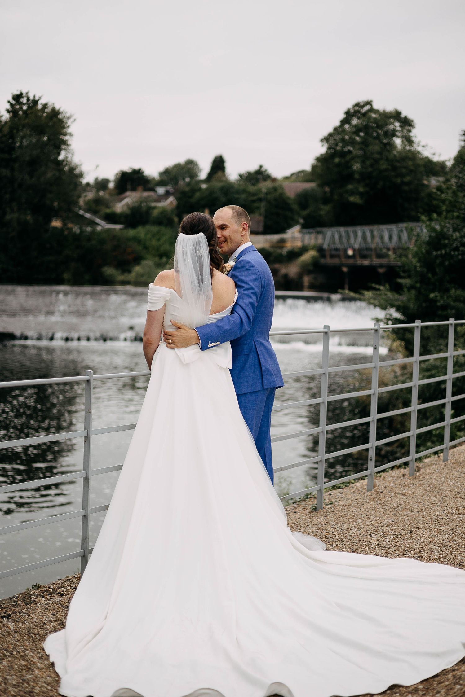 A bride and groom embrace near a riverside railing, she in a white gown with veil, he in a blue suit.