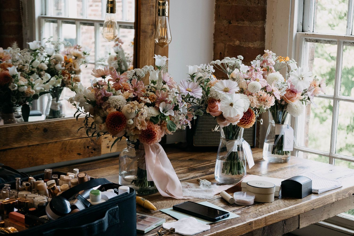 Elegant floral arrangements in glass vases with pink and white blooms displayed on a rustic wooden table near bright windows.