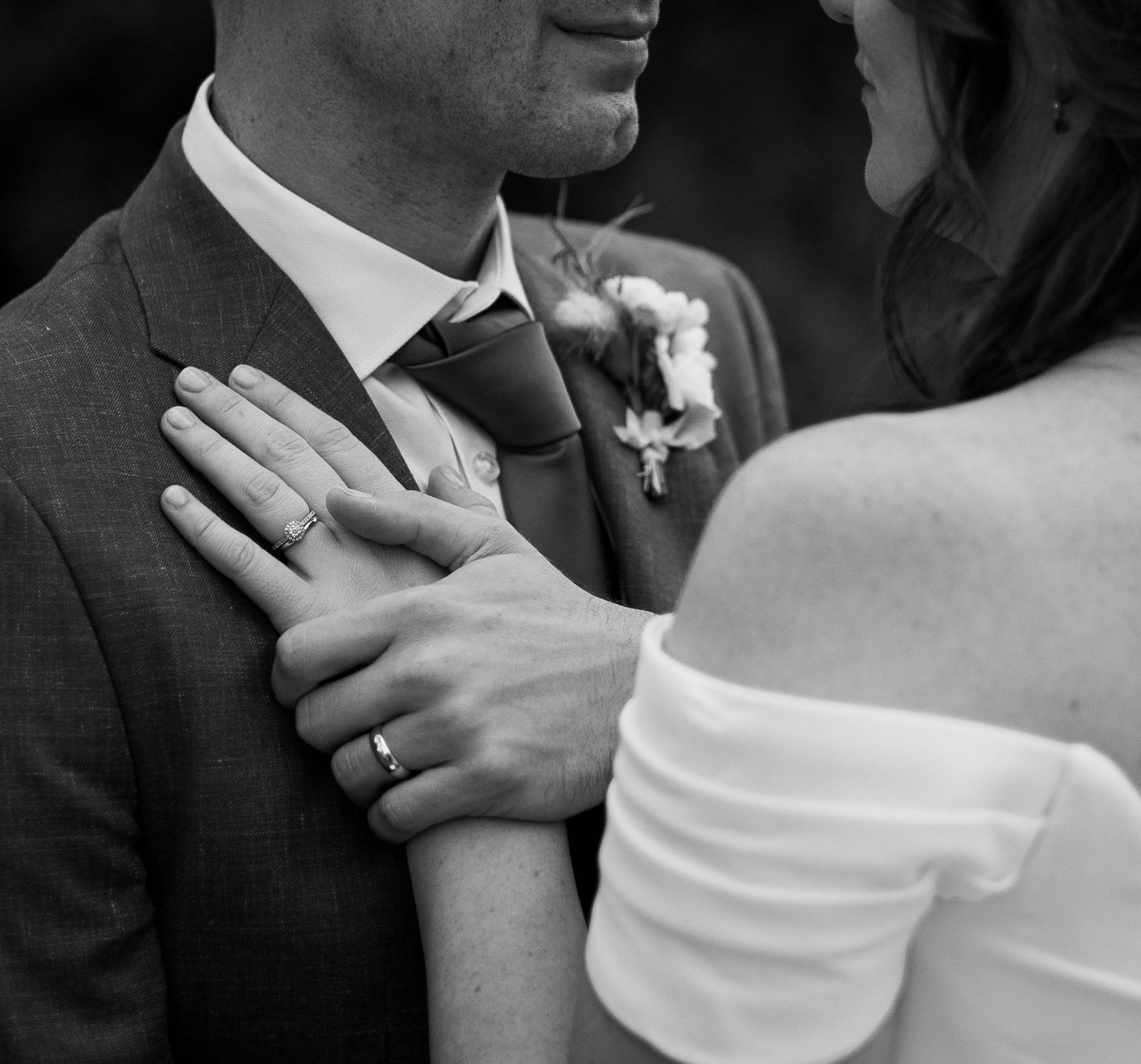 Black and white wedding photo of bride and groom embracing, showing wedding rings and boutonniere.