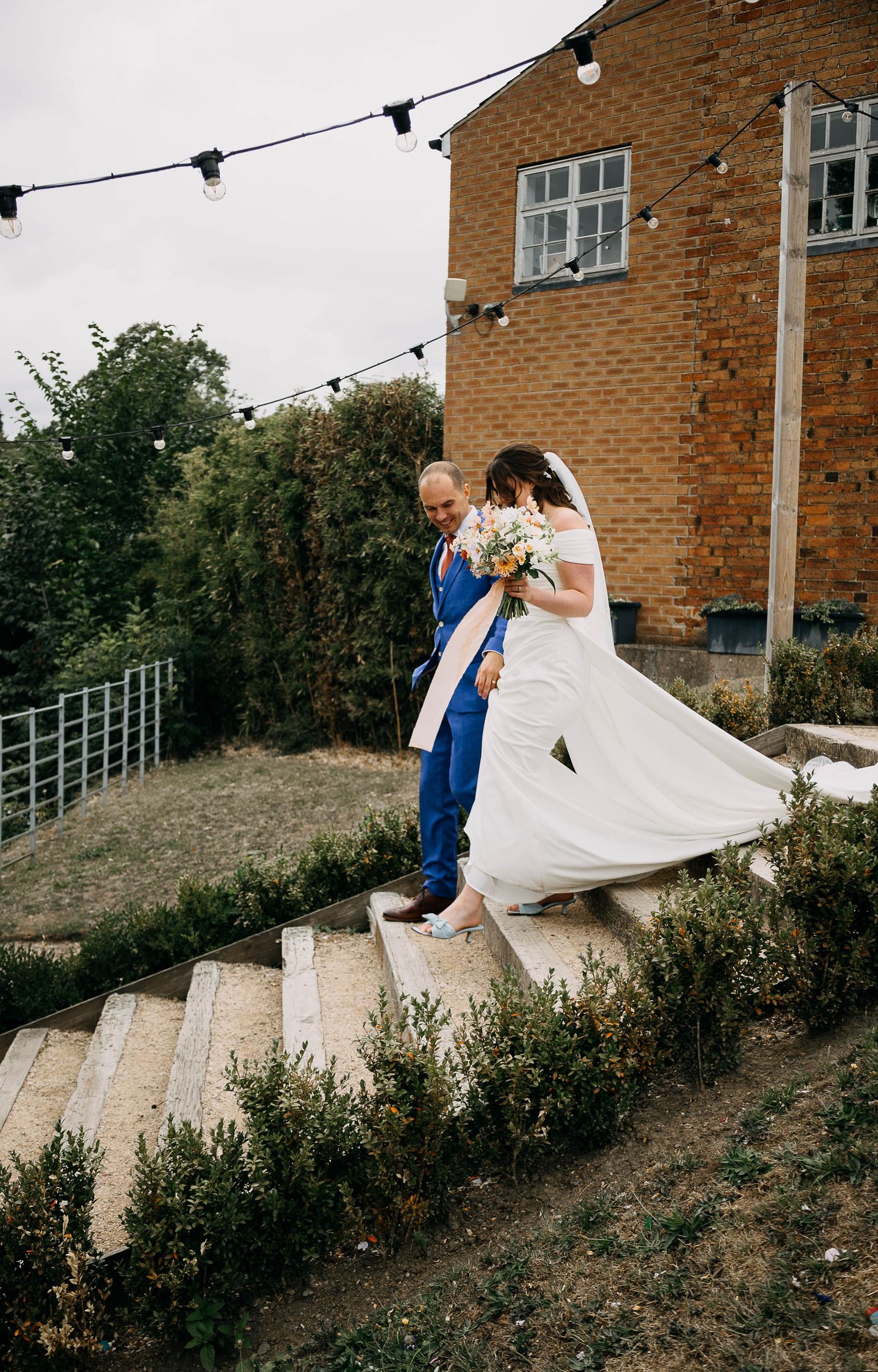 Bride in white gown and groom in blue suit walking down stone steps at outdoor wedding venue with string lights.