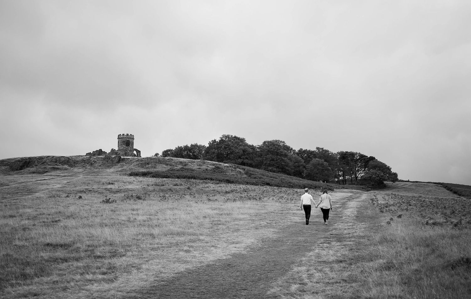 Two people walk along a rural path toward a hilltop tower in a moody black and white landscape.