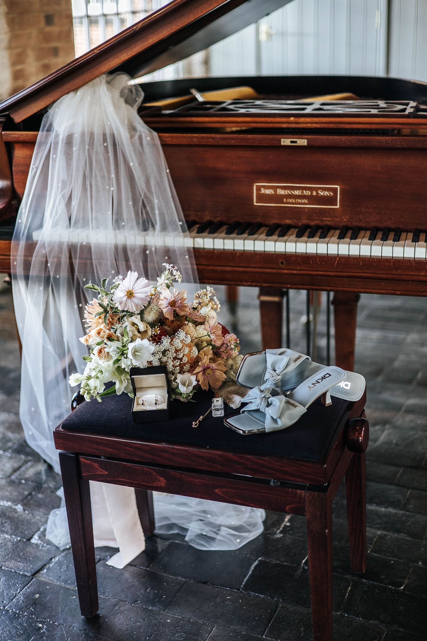 Wedding bouquet, gray heels, and ring box on piano bench with bridal veil draped over vintage grand piano.
