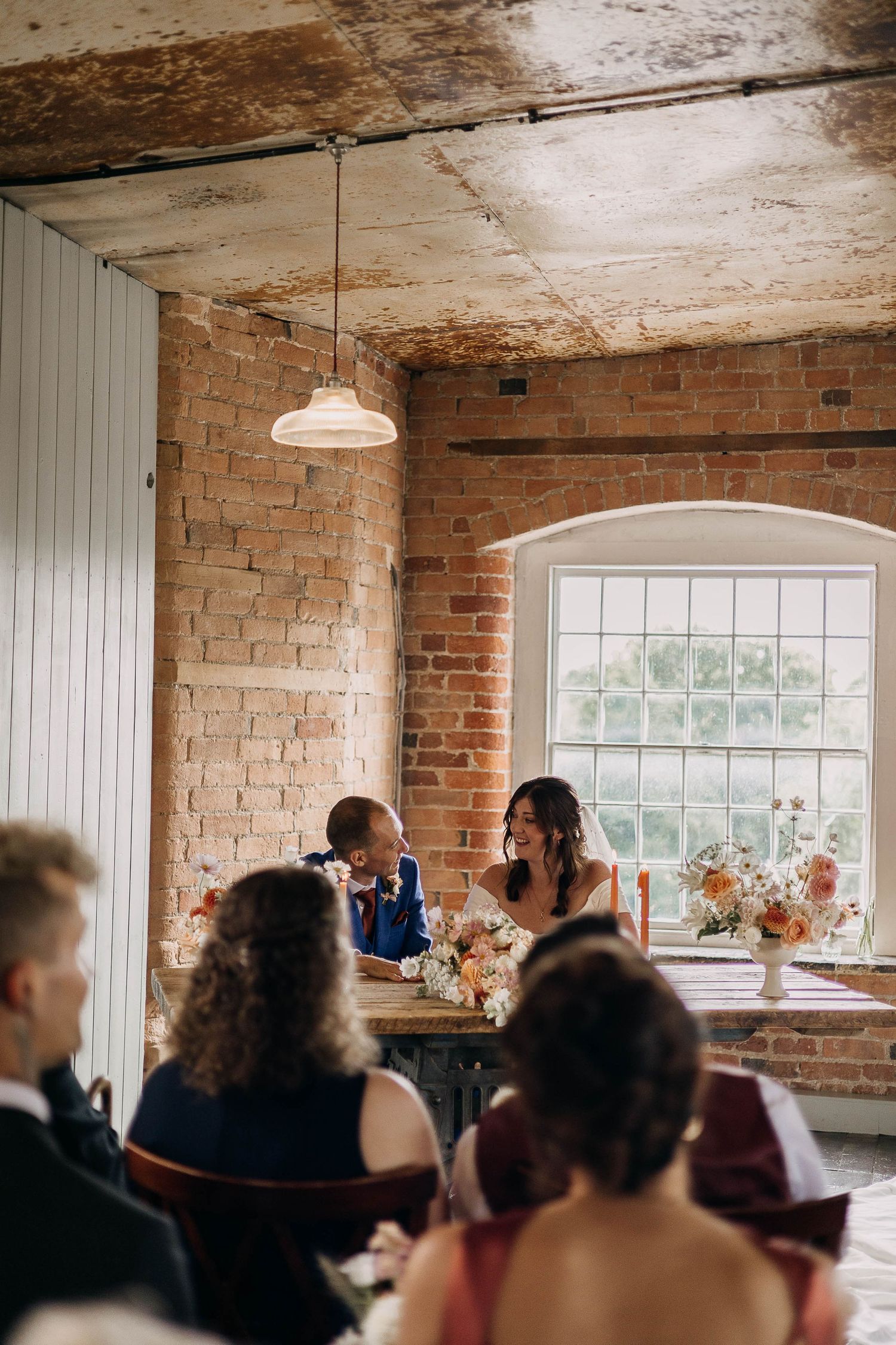 Bride and groom laughing at sweetheart table in rustic brick venue with floral centerpieces and guests seated nearby.