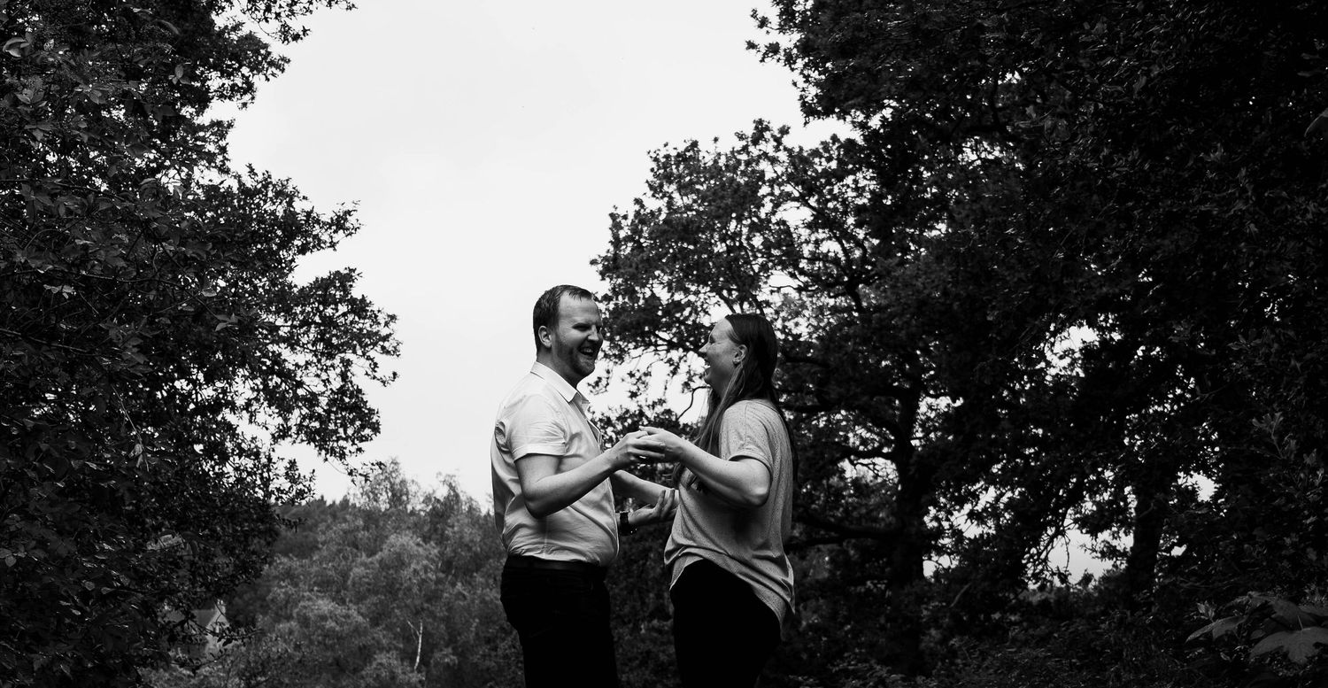 Black and white photo of a couple standing together among lush trees, sharing a romantic moment outdoors.