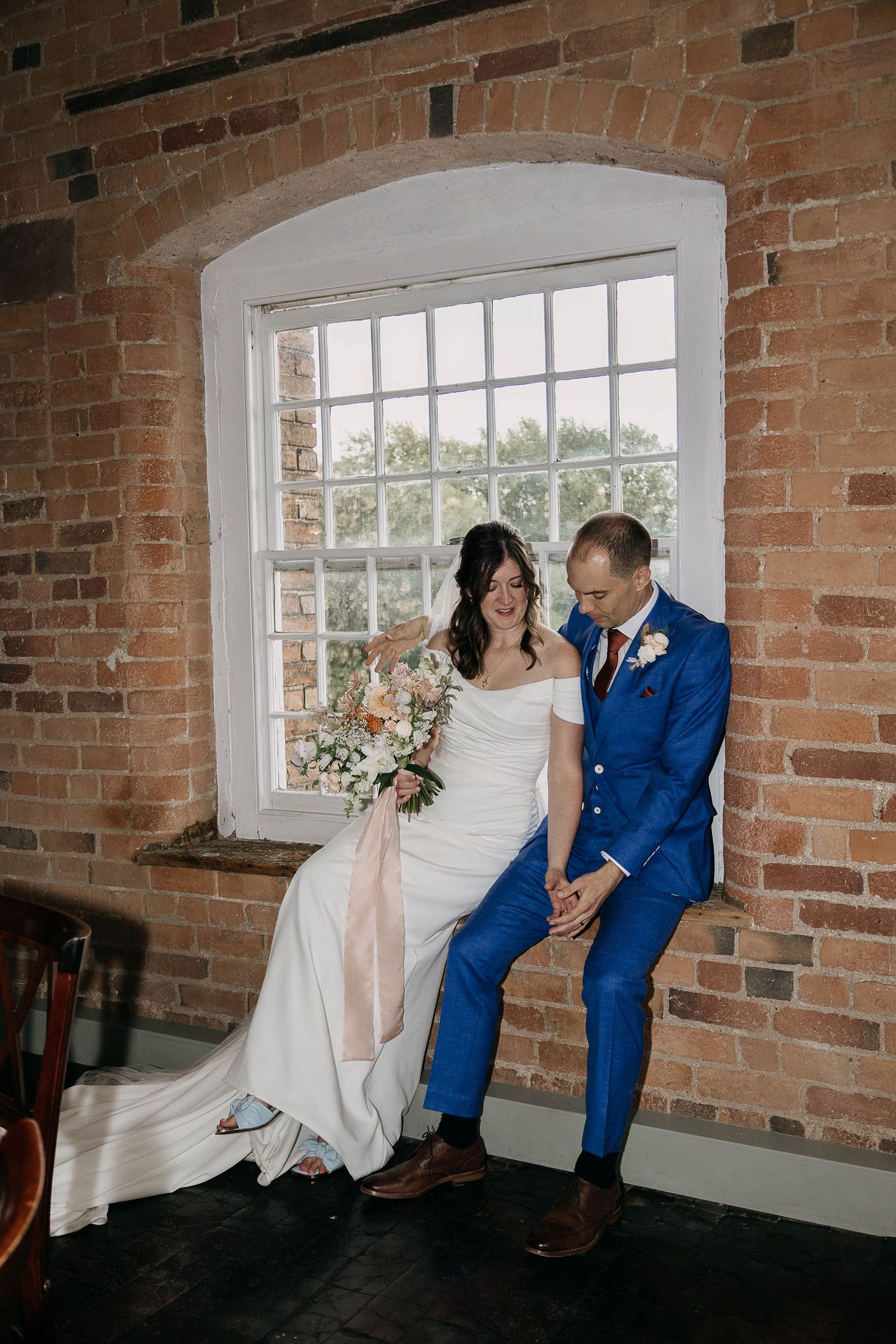 Bride and groom sit together by a brick wall window, she holds a pastel bouquet, he wears a bold blue suit.