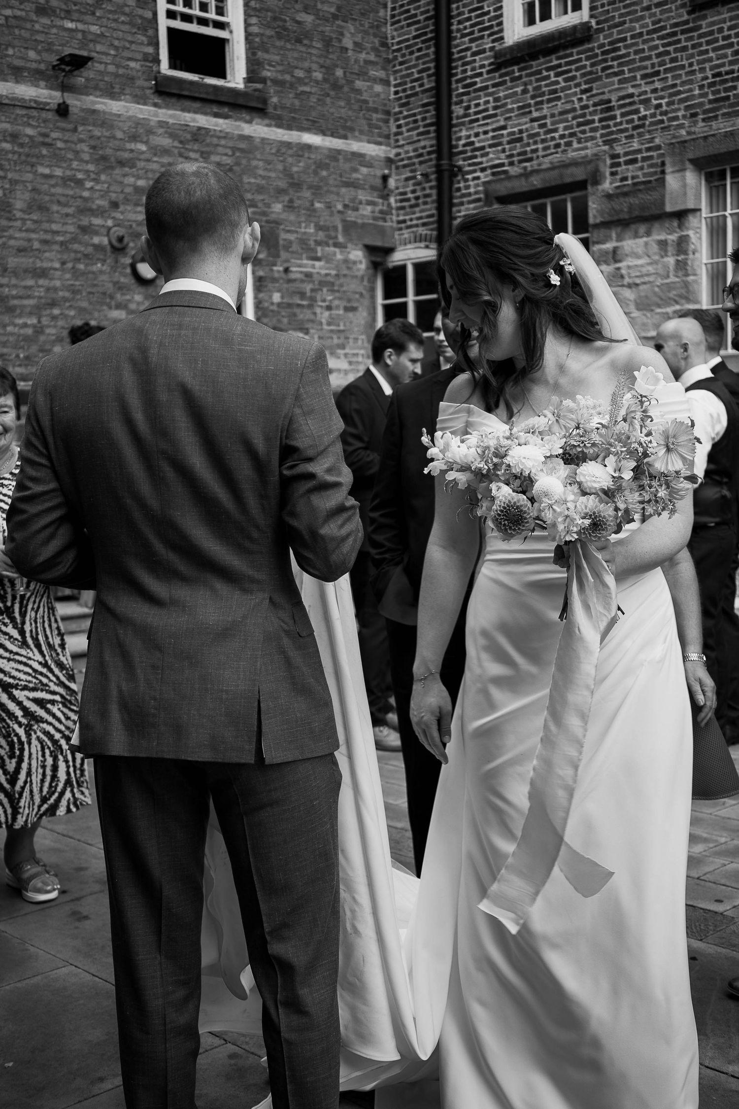 Black and white photo of a bride holding a floral bouquet standing beside a groom outside a brick building.