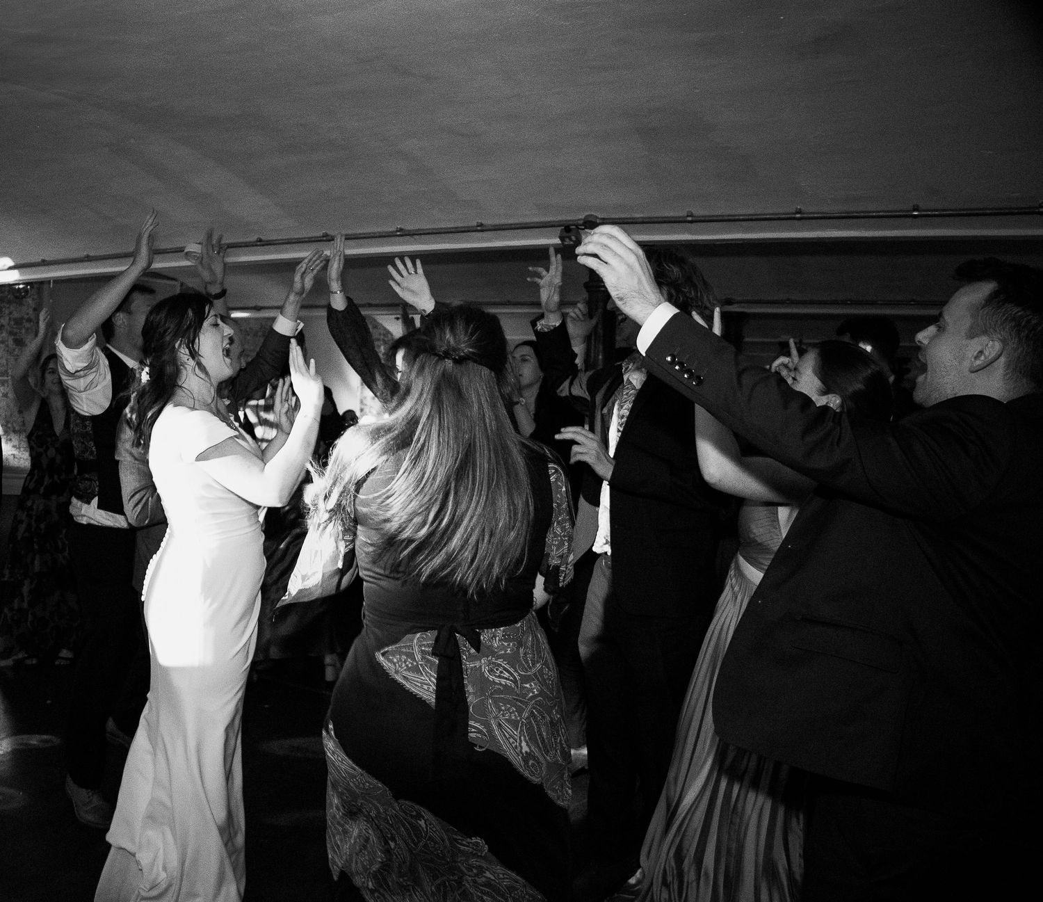 Black and white photo of wedding guests dancing joyfully with hands raised at a lively reception party.