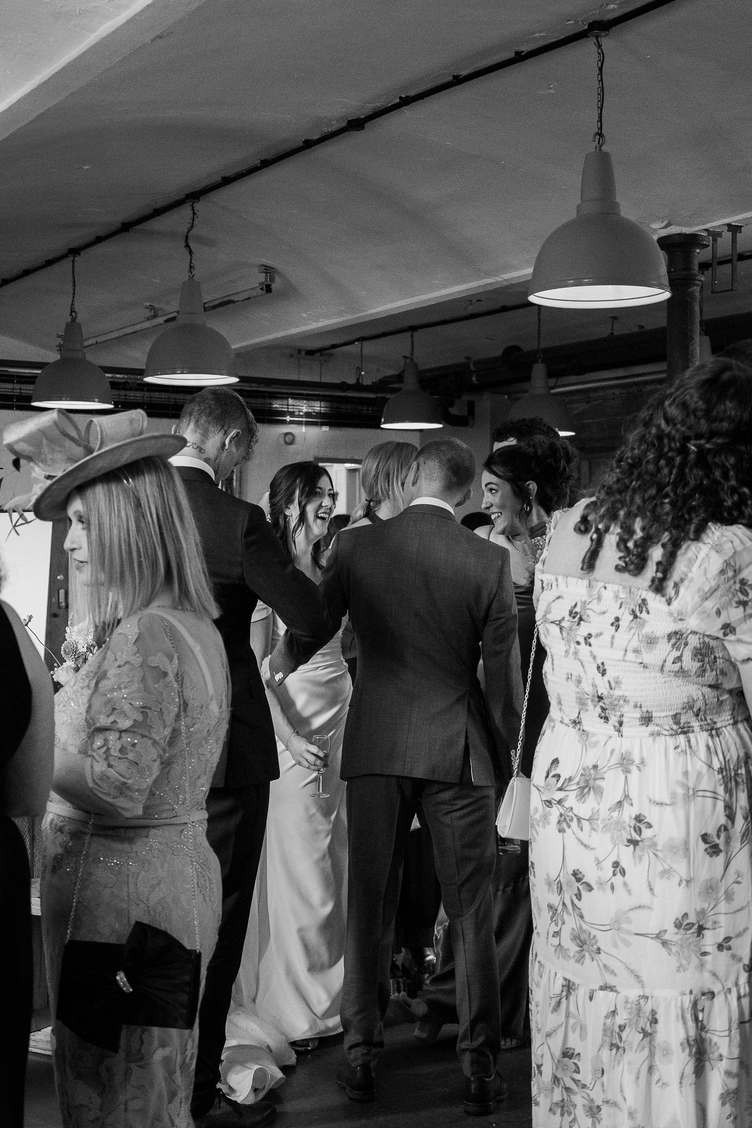 Black and white photo of wedding guests mingling indoors, bride in white gown laughing with groom in suit.