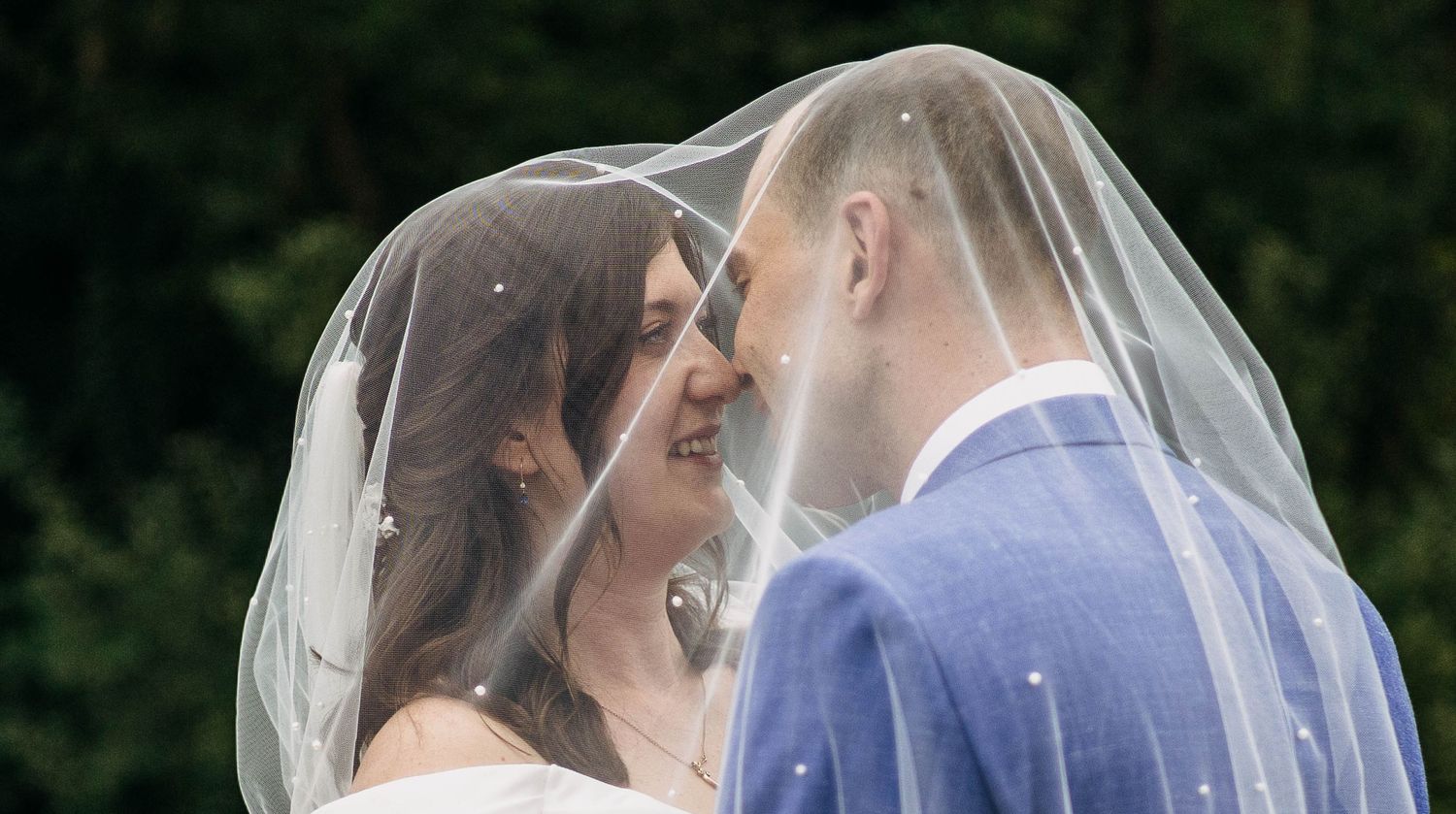 A bride and groom share a romantic moment under a sheer veil, smiling at each other on their wedding day.
