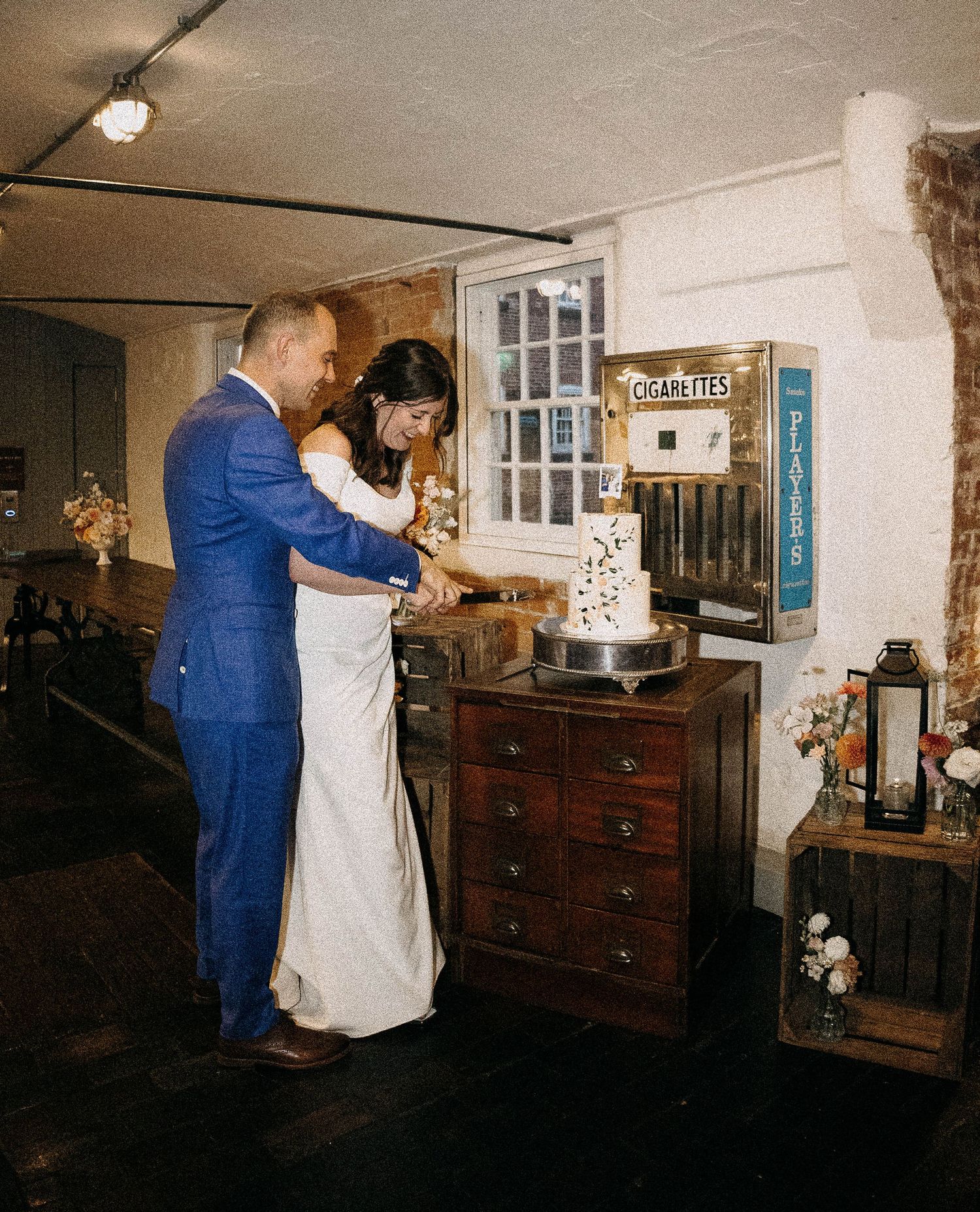 Bride and groom cutting their wedding cake in a rustic venue beside a vintage cigarette machine.