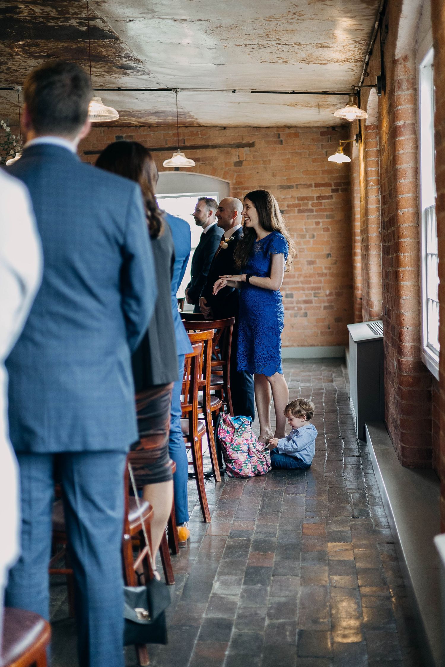 Guests mingling in a rustic brick venue during a formal event, with a child sitting on the floor beside a woman in blue.