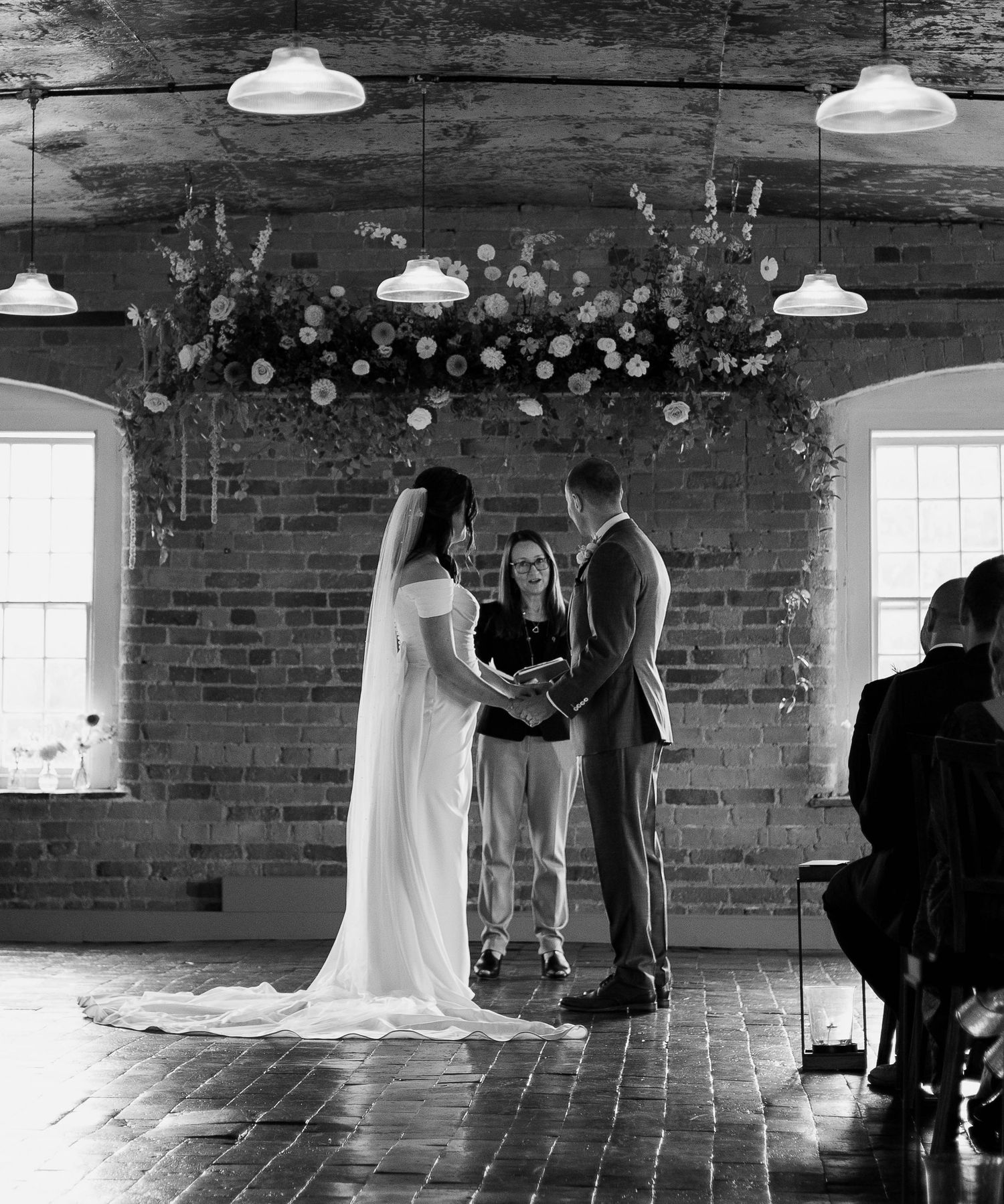 Black and white photo of a bride and groom exchanging vows indoors at a rustic brick venue with floral arch decor.