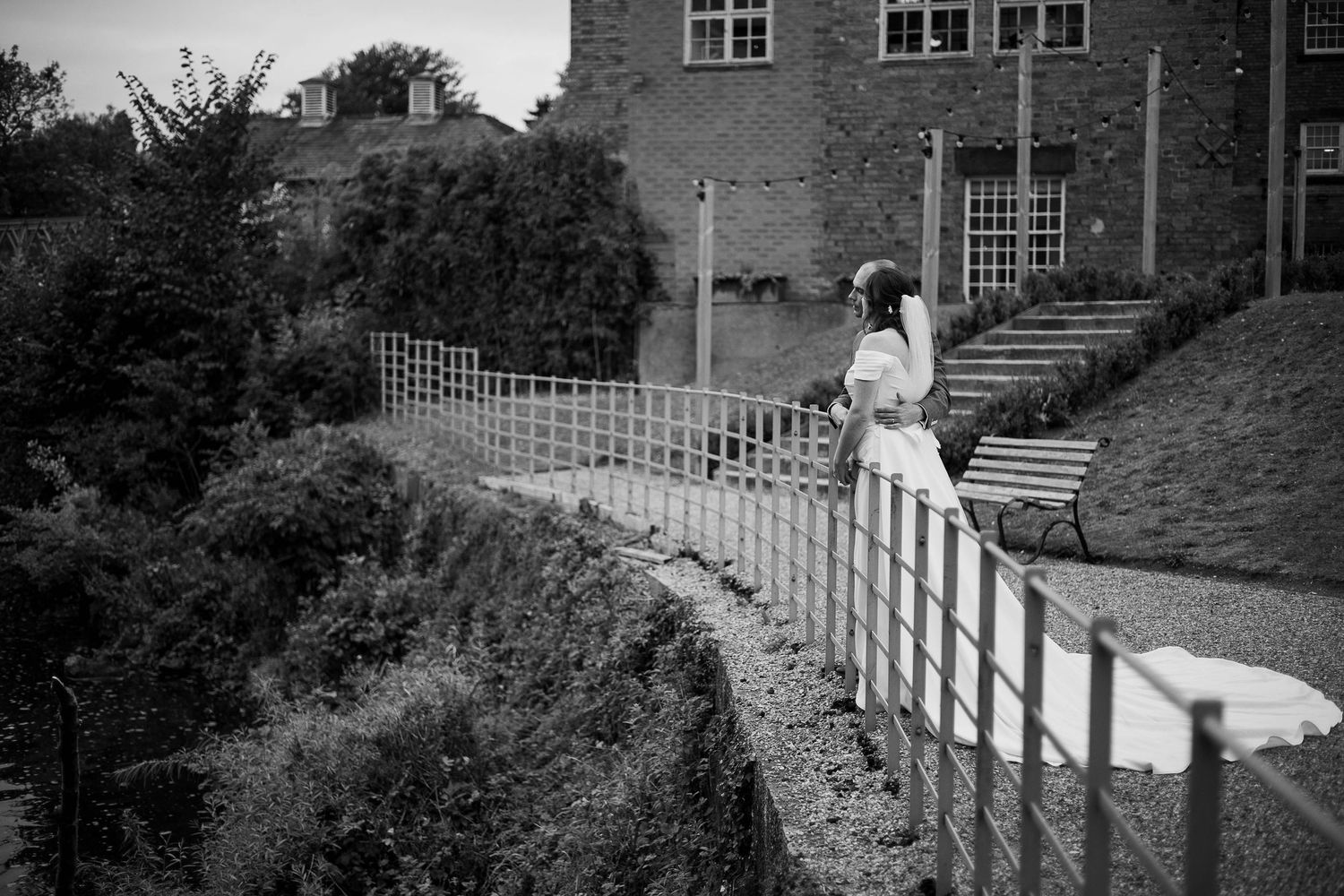 Black and white photo of a bride standing by a white fence outside a brick building surrounded by lush greenery.