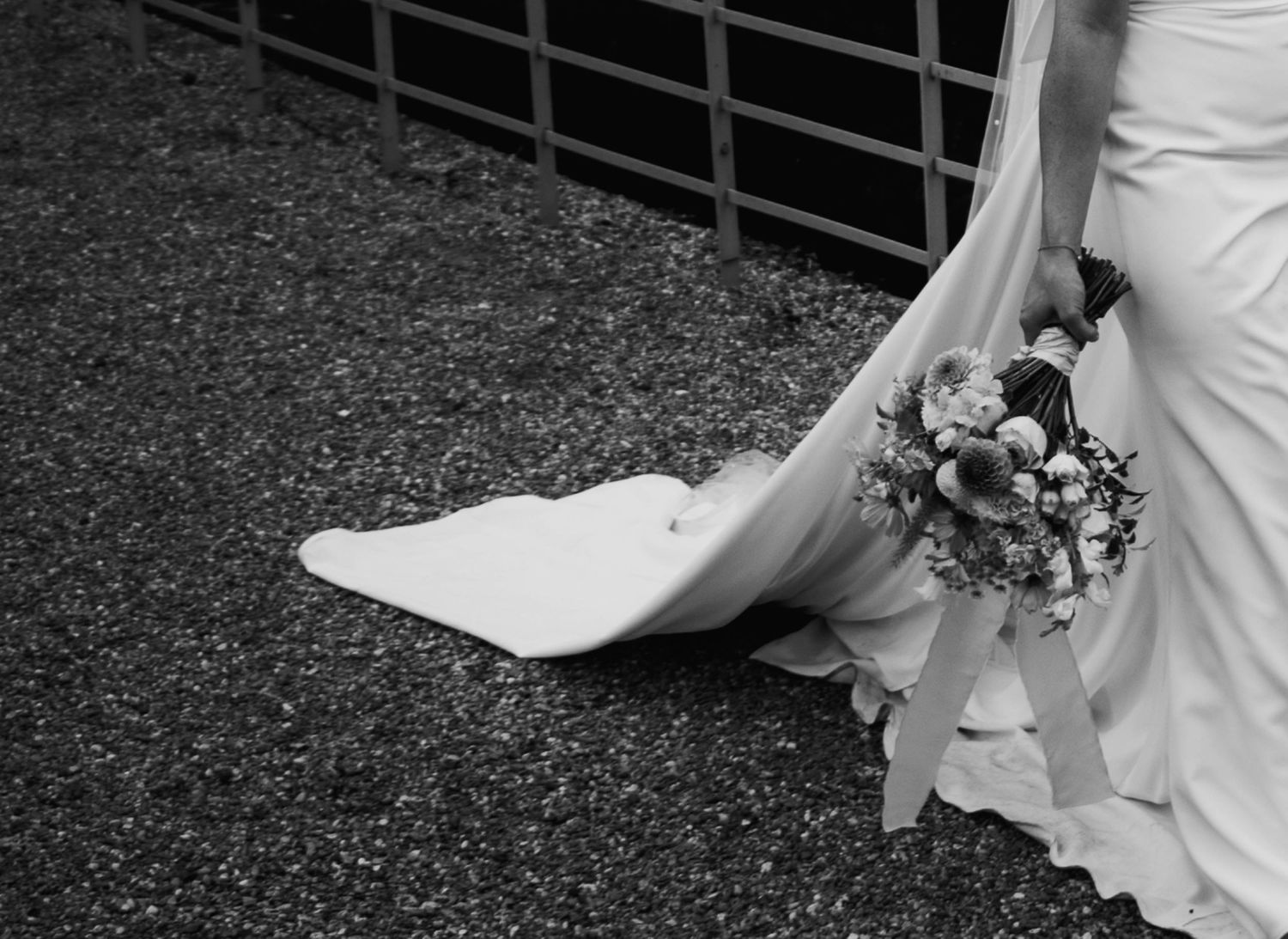 Black and white photo of a bride holding a floral bouquet, with her wedding dress train spread on gravel ground.