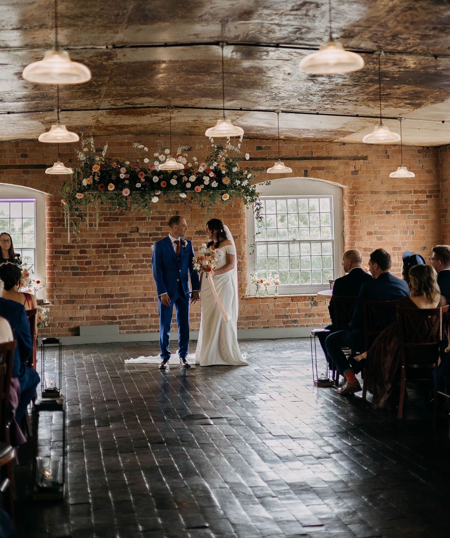 Couple exchanges vows at rustic brick wedding venue with floral arch, pendant lights, and seated guests.