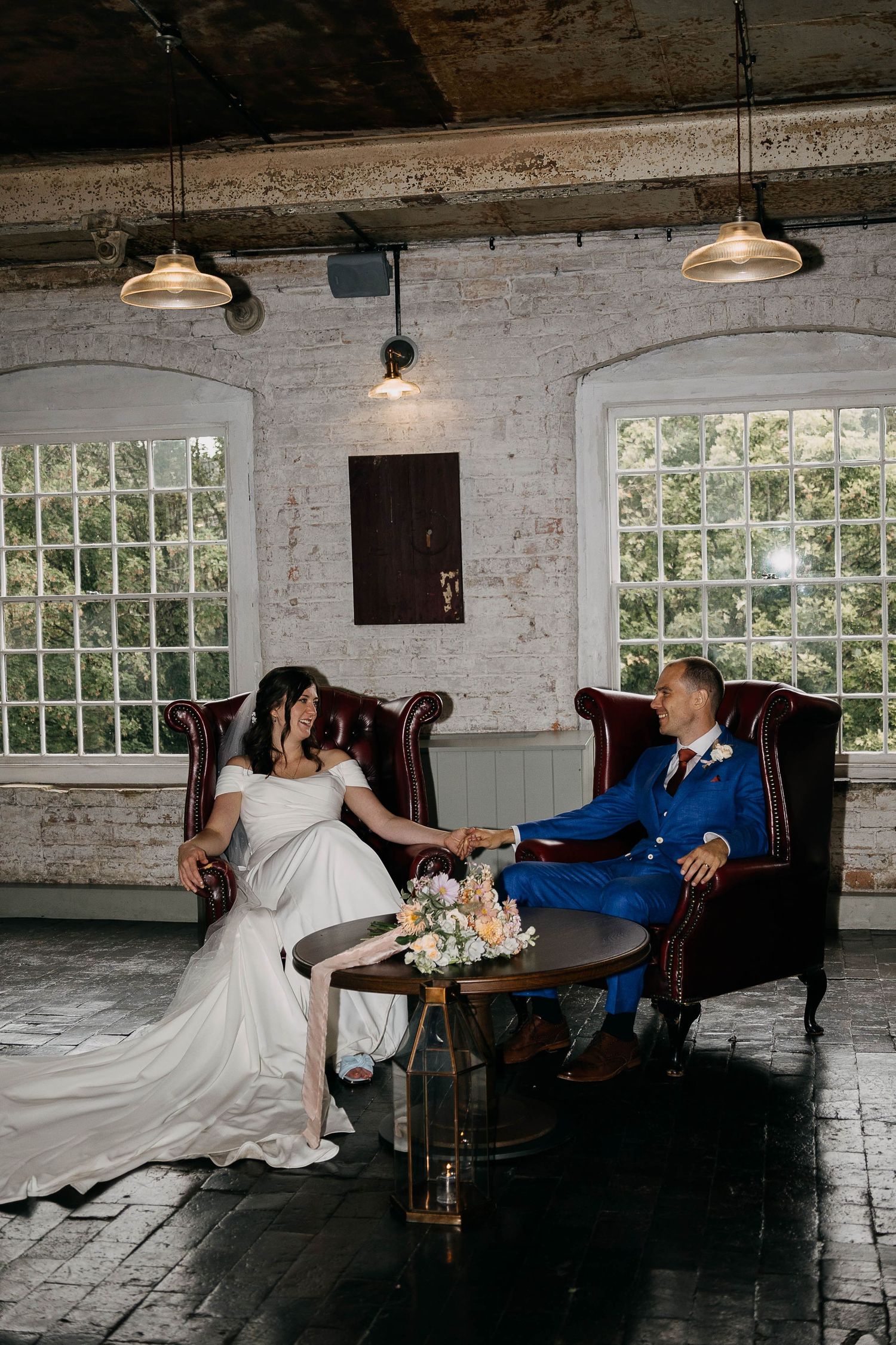 Bride and groom sitting on vintage leather chairs holding hands at an industrial chic wedding venue with white brick walls.
