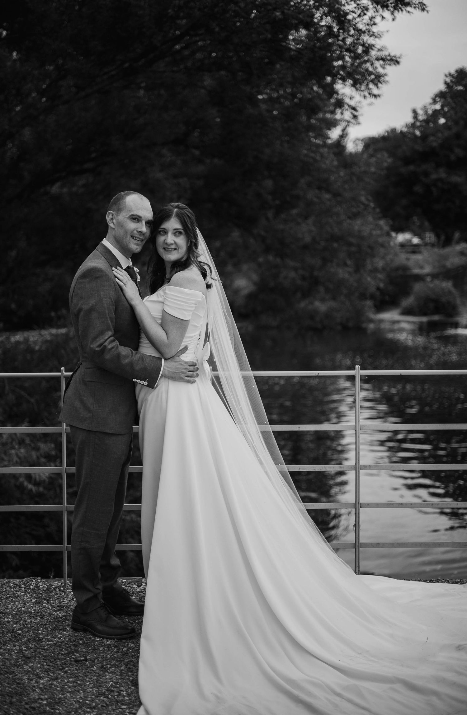 Black and white wedding photo of bride in flowing gown with veil and groom embracing near a serene waterfront.