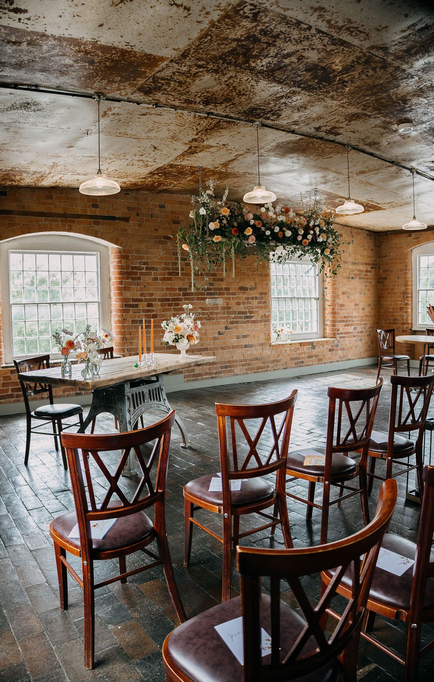 Rustic wedding venue with exposed brick walls, wooden chairs, round tables, and floral ceiling installation with hanging greenery.