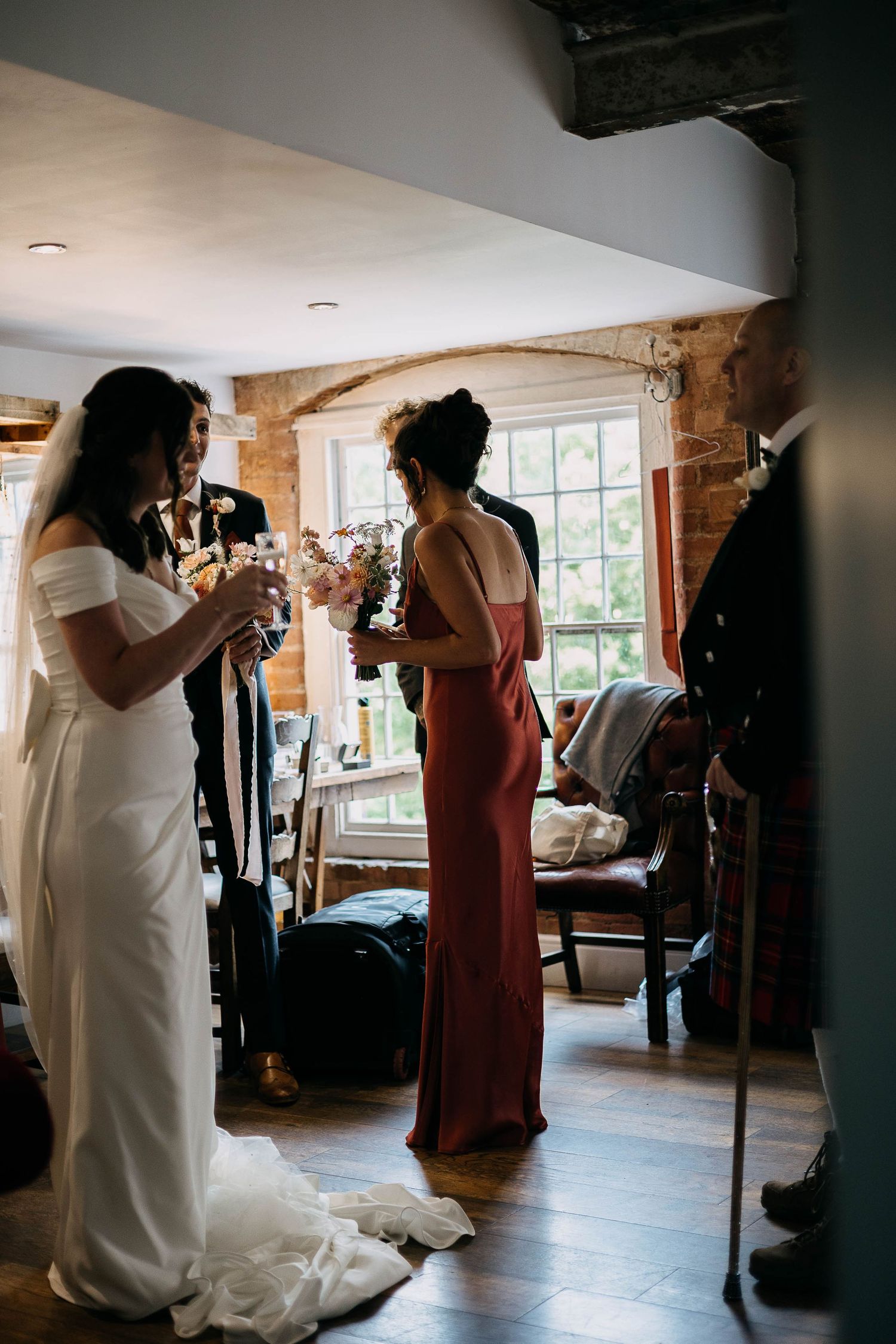 Bride in white gown and bridesmaid in red dress holding bouquet toast with guests in rustic brick venue.