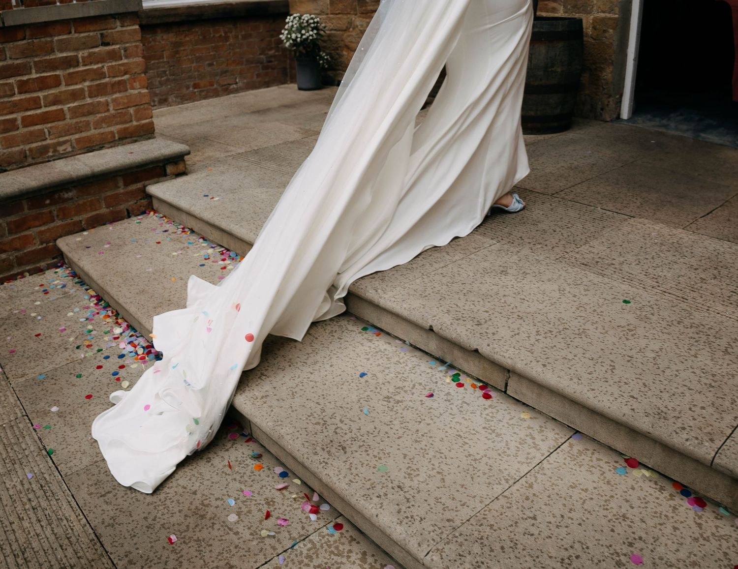 A bride's white wedding dress train draped over stone steps covered in colorful confetti outside a brick building.