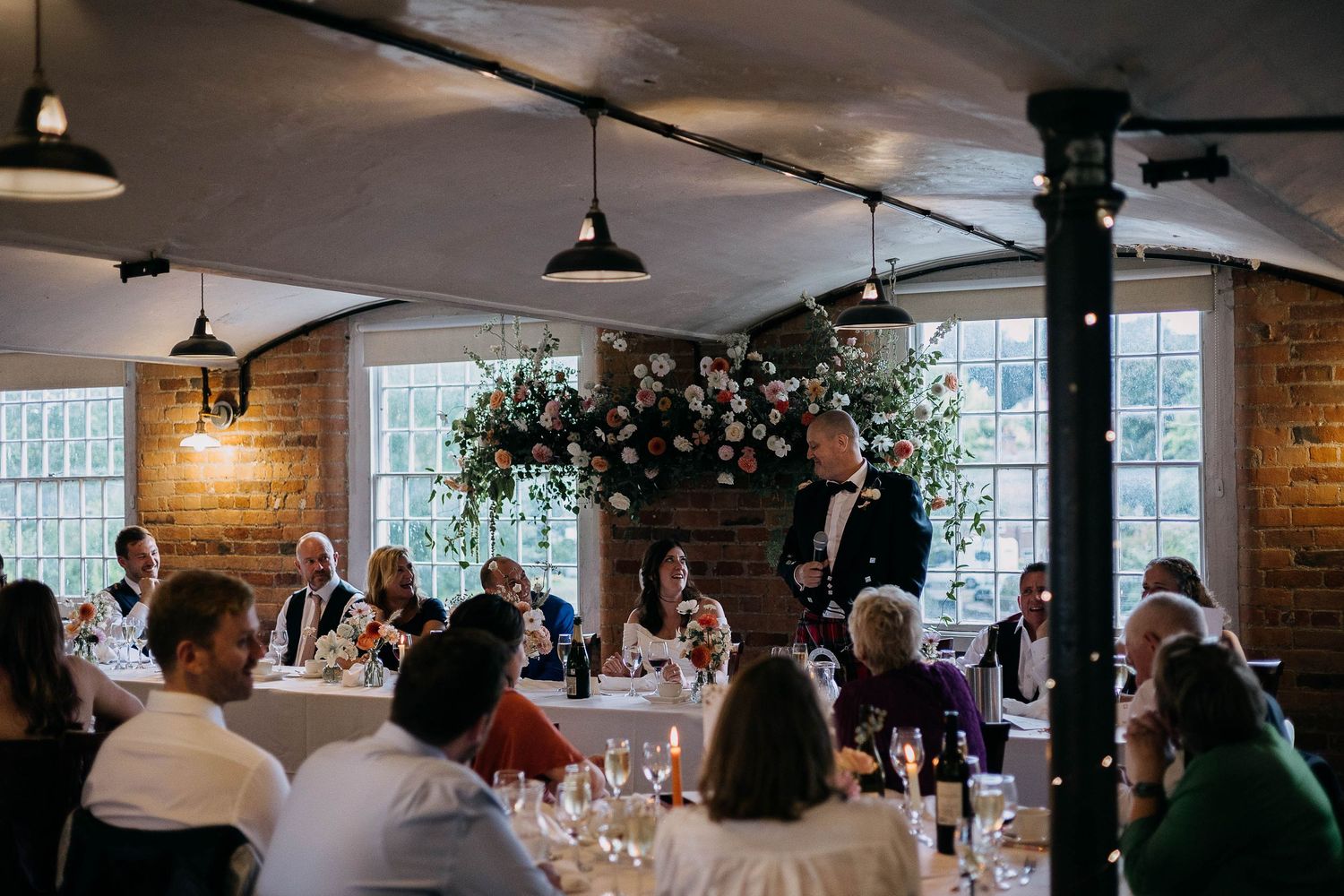 Elegant wedding reception with guests seated at round tables, floral arch backdrop, and groom giving a speech.