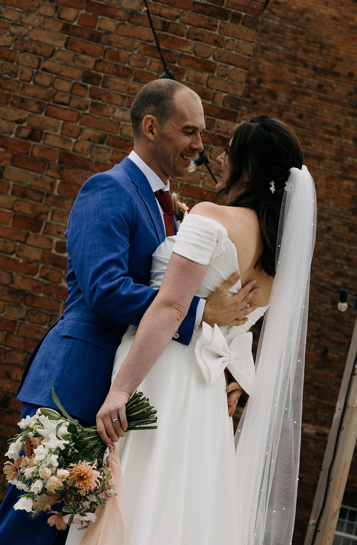 A groom in a blue suit embraces his bride in a white off-shoulder gown and veil, holding a bouquet indoors.