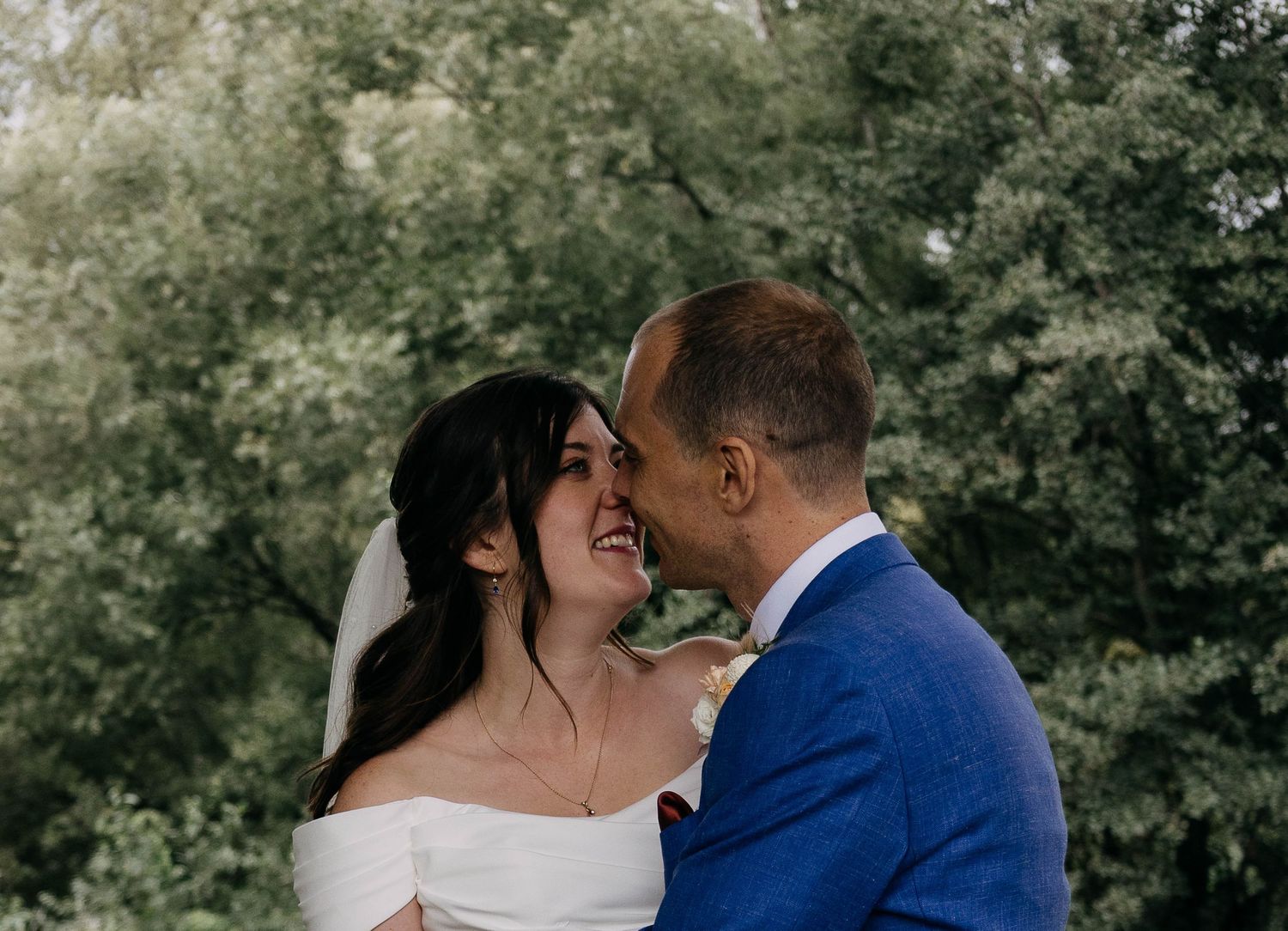 A bride and groom share a romantic moment outdoors, smiling at each other with lush green trees in the background.
