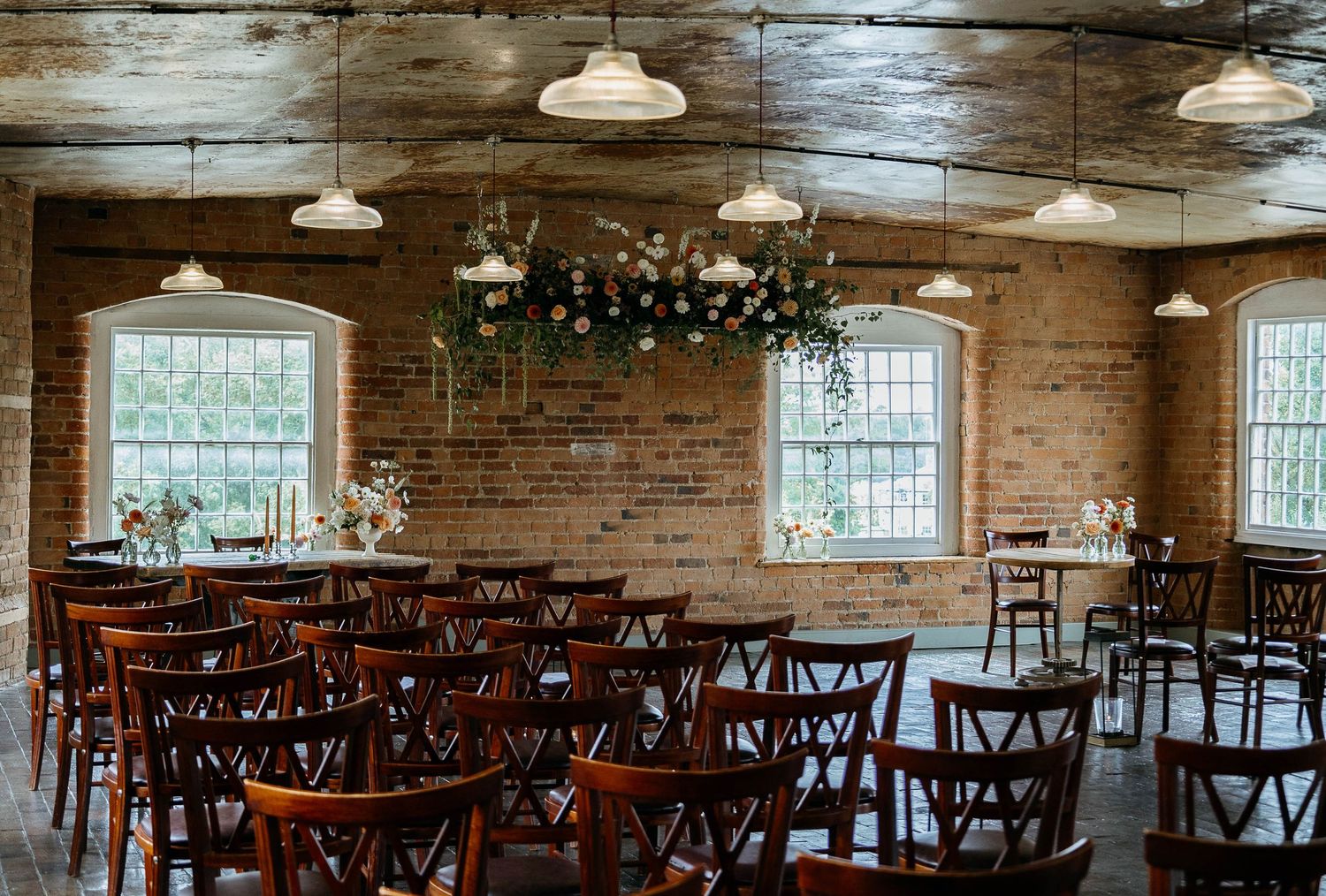 Rustic brick wedding venue with wooden chairs, floral chandelier, and industrial pendant lights.