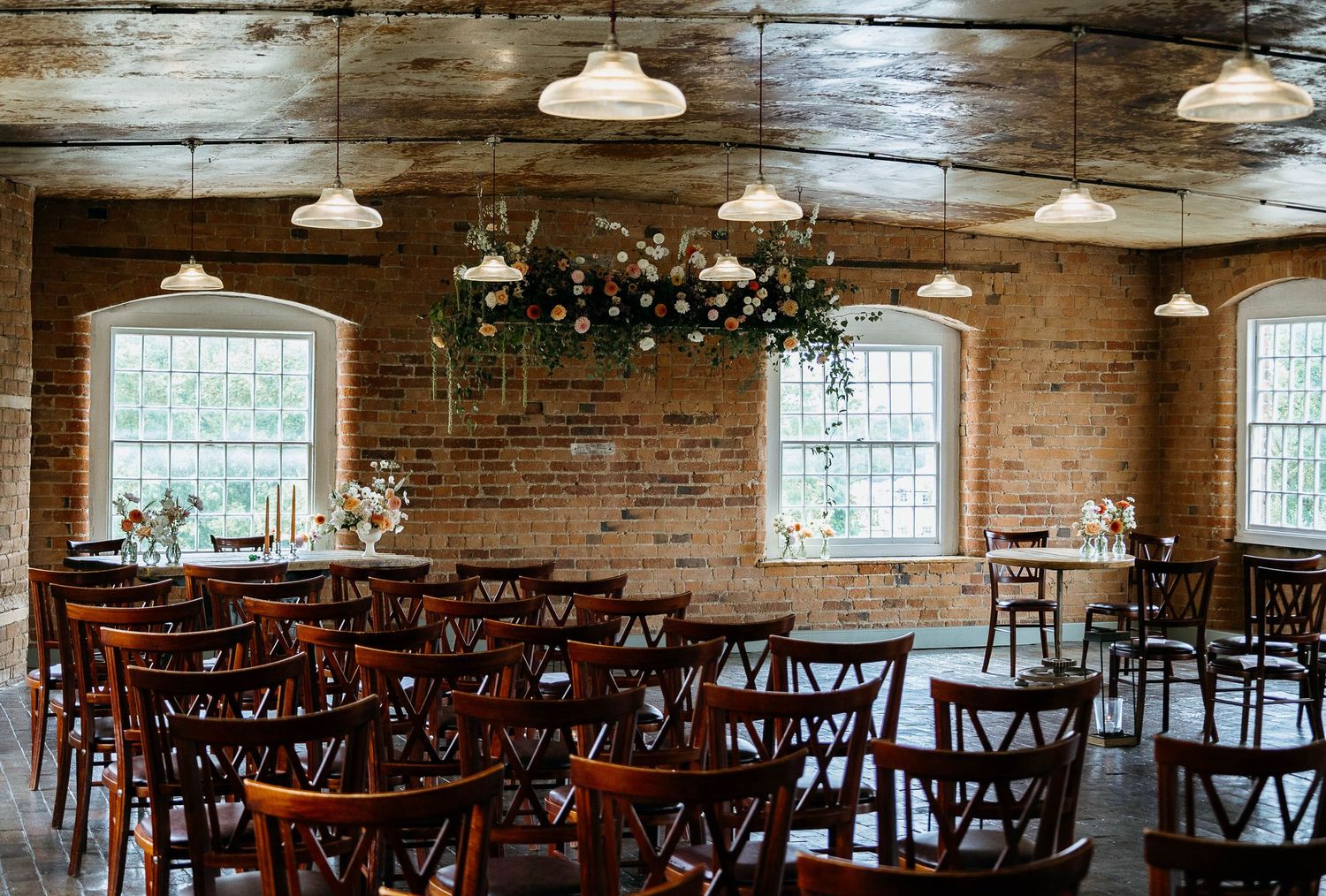 Rustic brick wedding venue with wooden chairs, floral chandelier, and industrial pendant lights creating an elegant ceremony space.