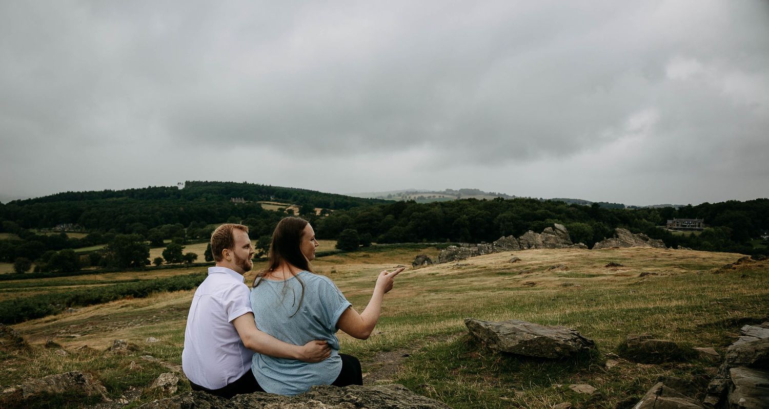 Couple sitting on rocks overlooking a vast green countryside landscape under a cloudy sky.