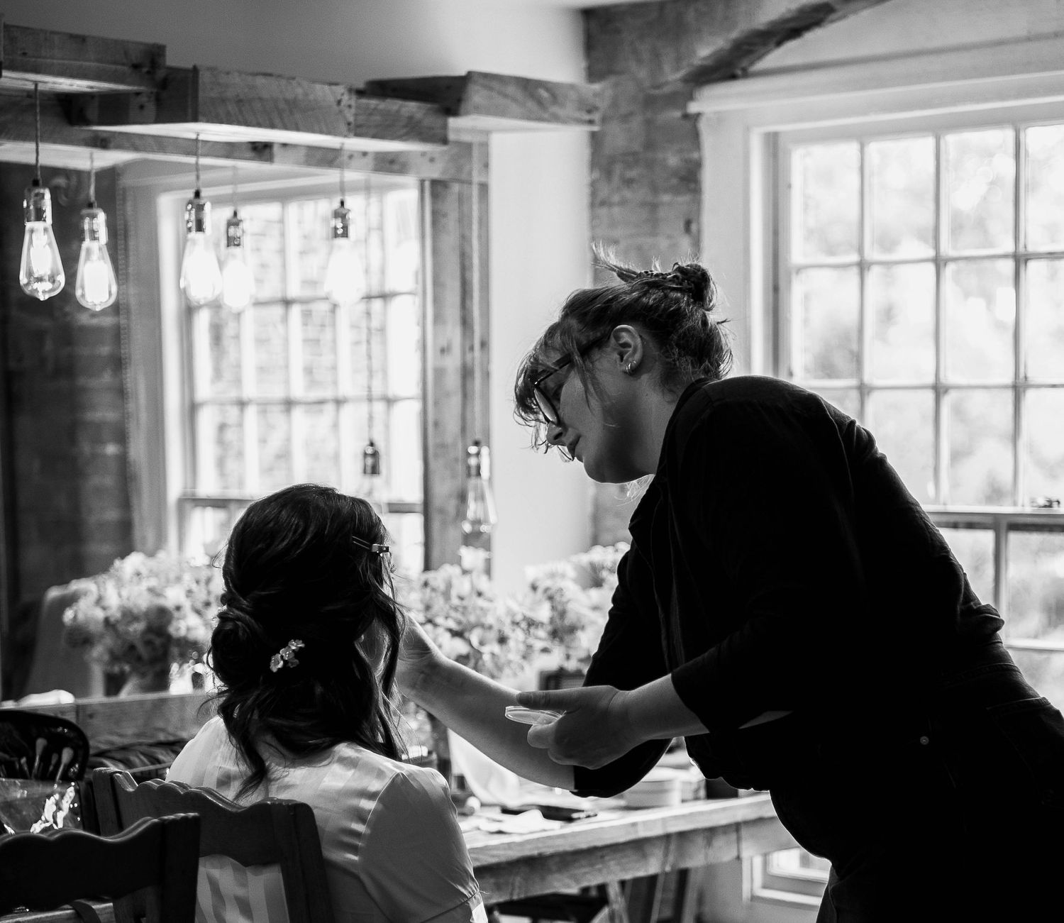 Black and white photo of a hairstylist working on a seated woman's hair in a rustic, warmly lit room.