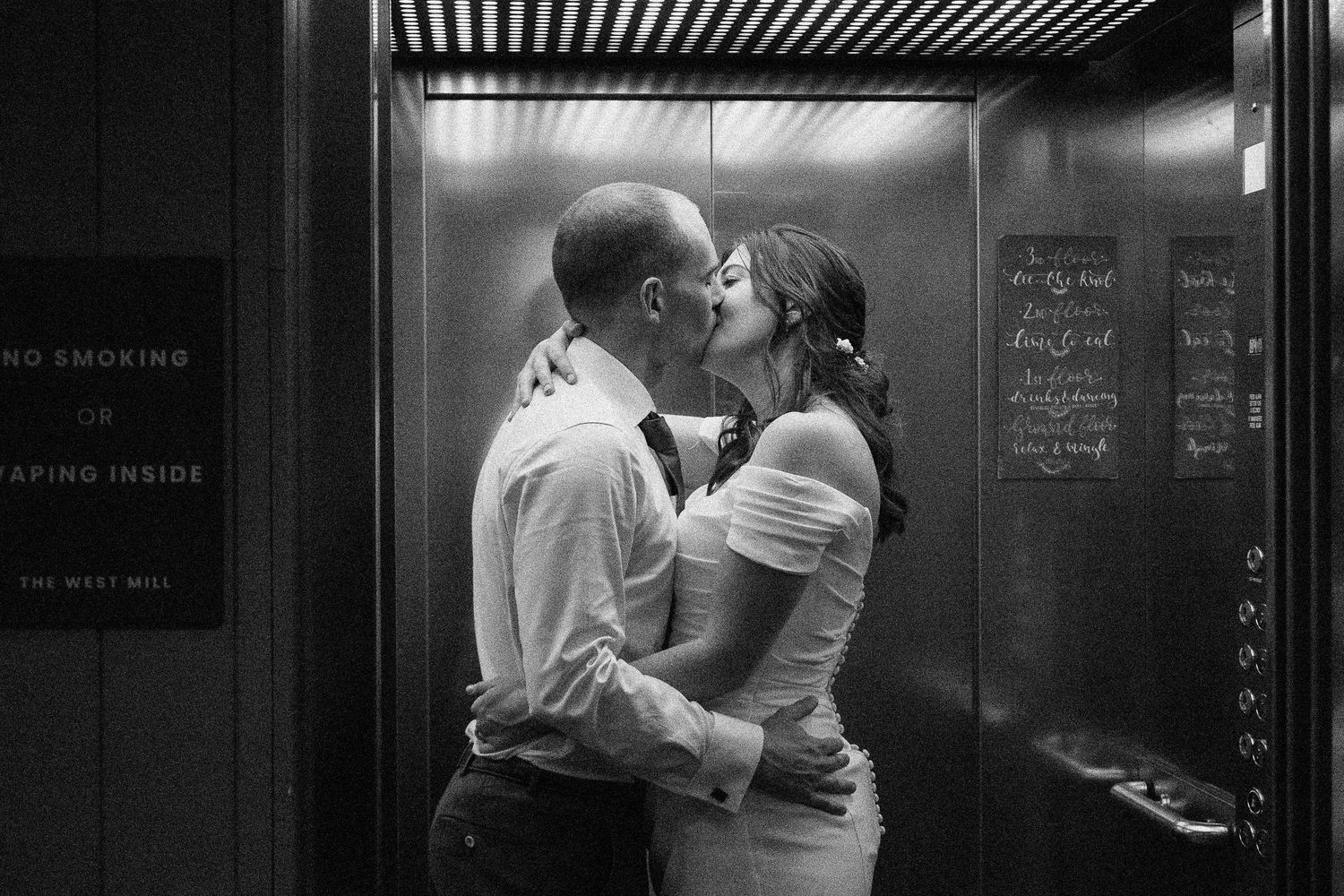 Black and white photo of a couple sharing a romantic kiss inside an elevator, the bride in a white dress.