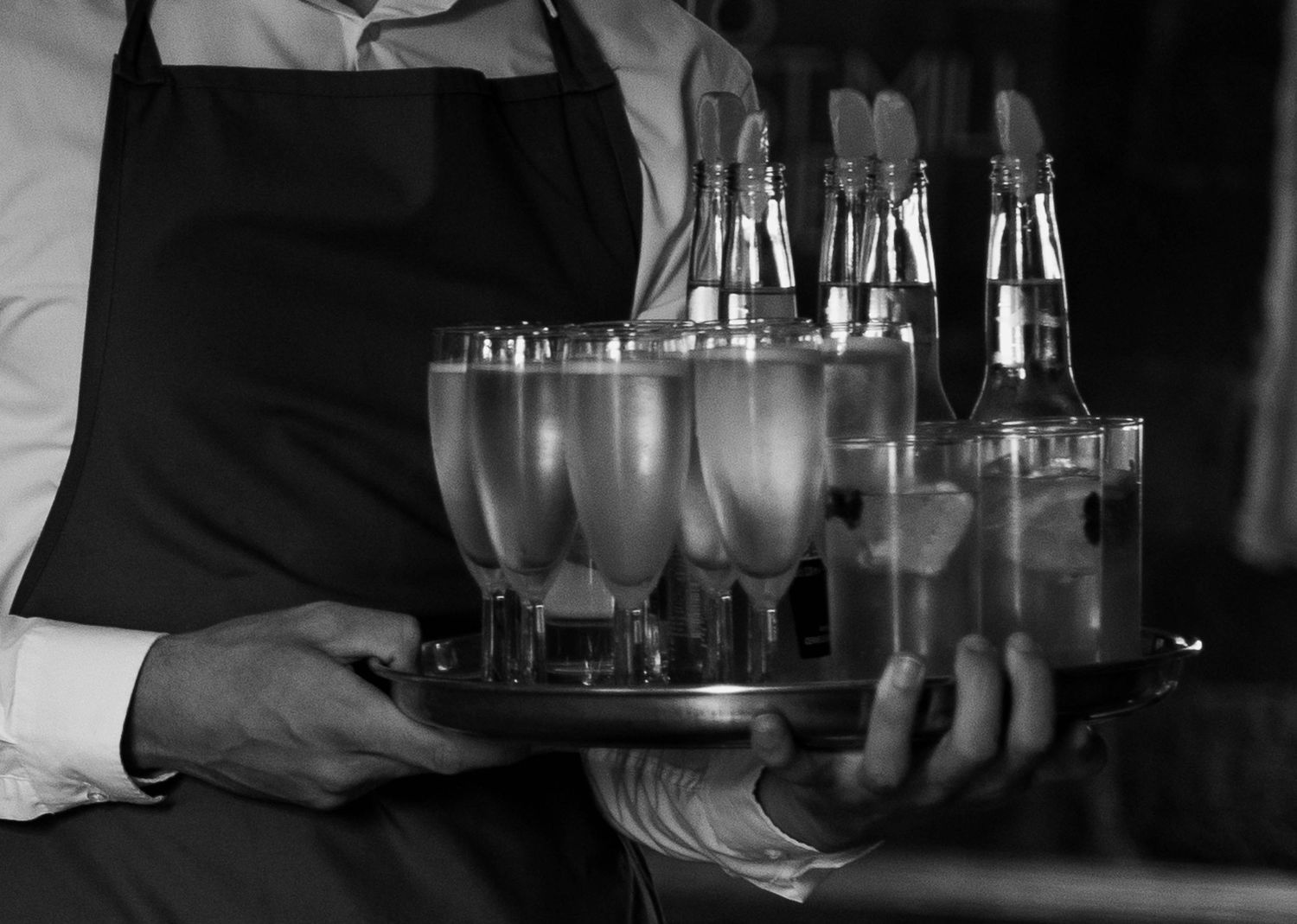 Waiter in black apron carrying a silver tray with champagne flutes and bottles at a formal event.