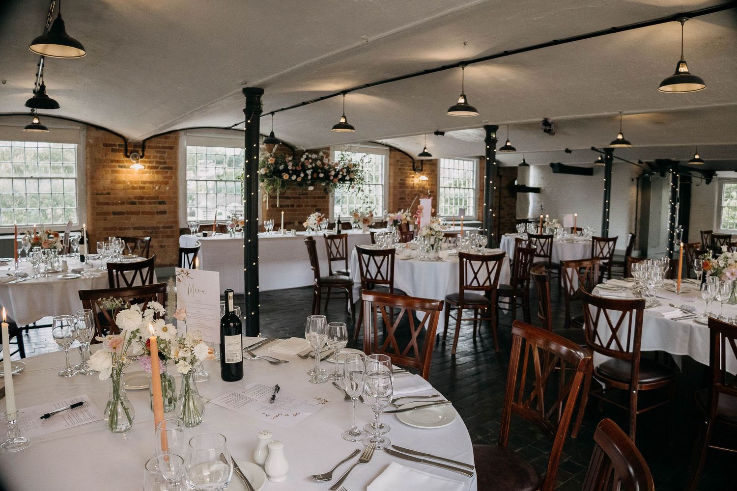 Elegant wedding reception venue with round white-clothed tables, wooden chairs, brick walls, and pendant lighting.