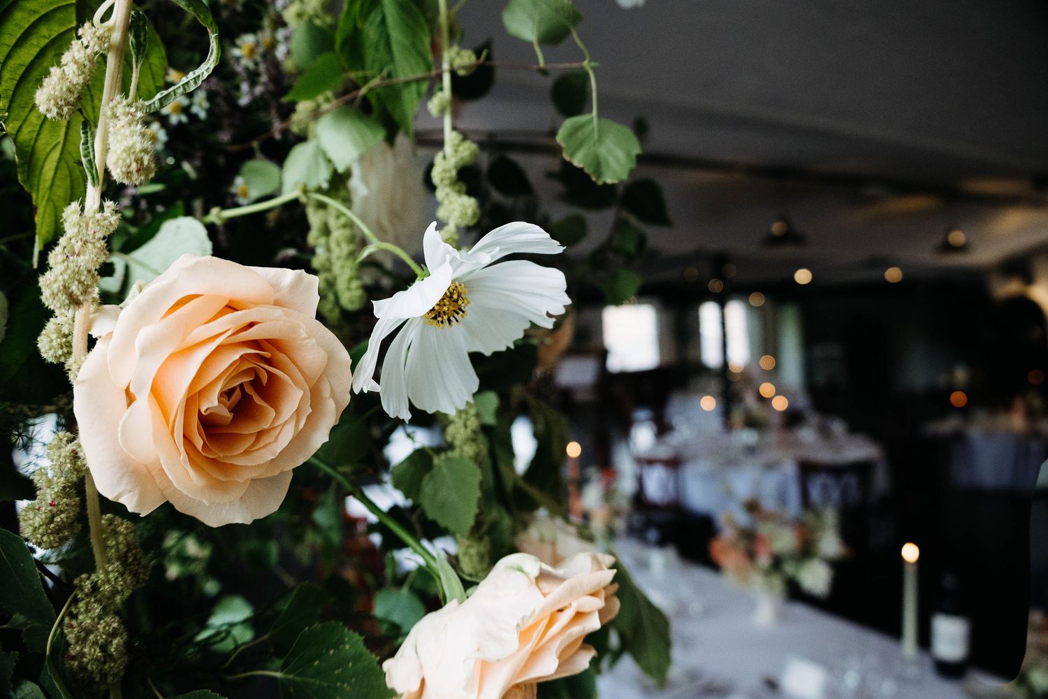 Elegant wedding reception centerpiece featuring peach roses and white cosmos flowers with lush greenery in a softly lit venue.